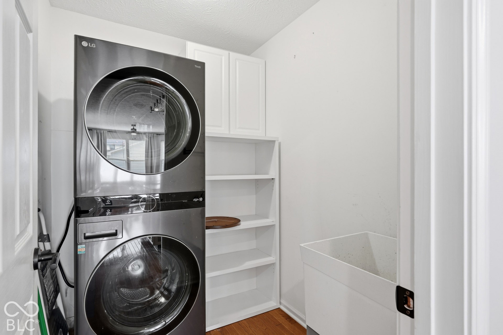 Stacked stainless steel laundry appliances, white upper cabinetry, open shelving, utility sink, and wood-finish flooring