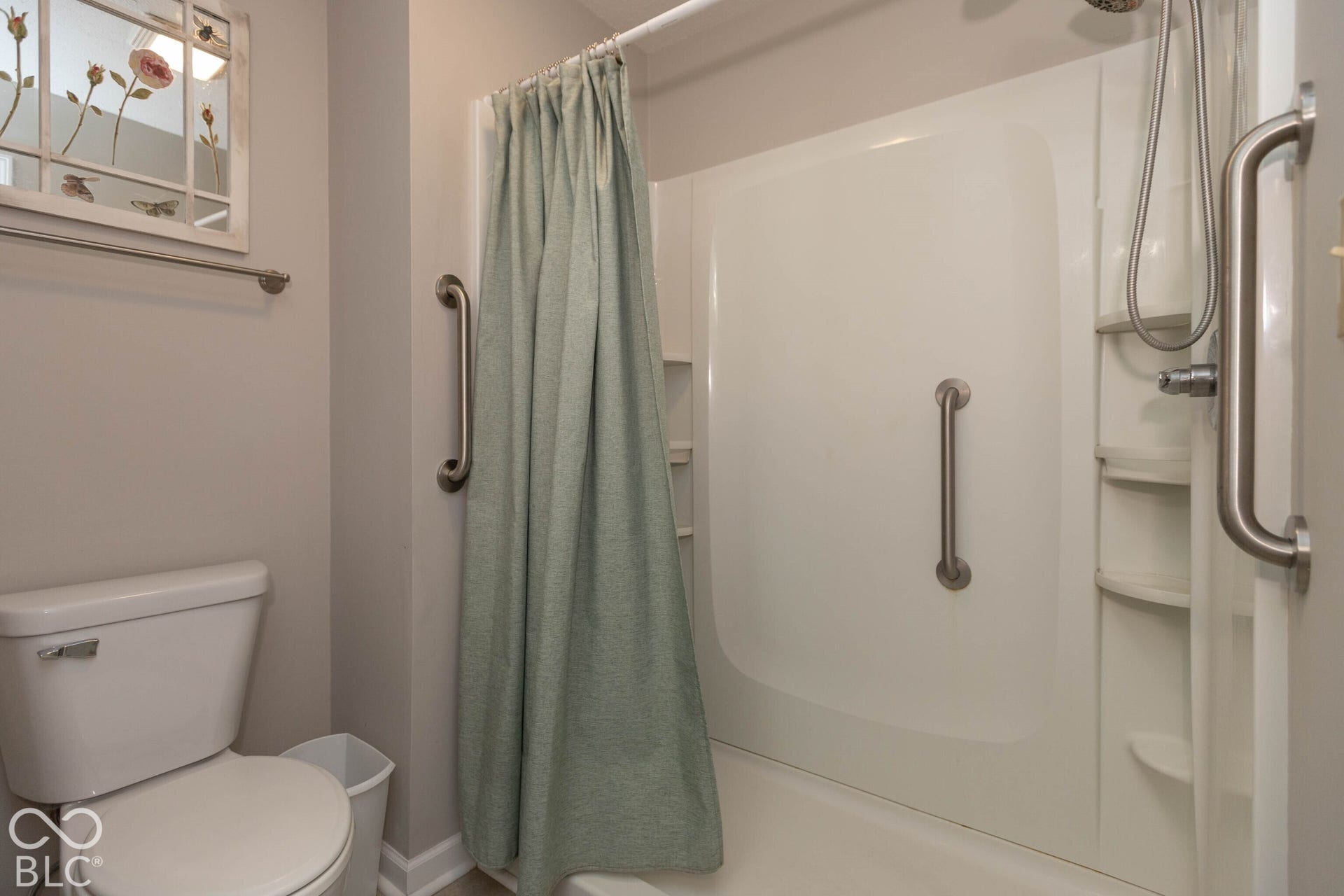 White shower enclosure with integrated shelving, multiple grab bars, and a wall-mounted shower head with a flexible hose