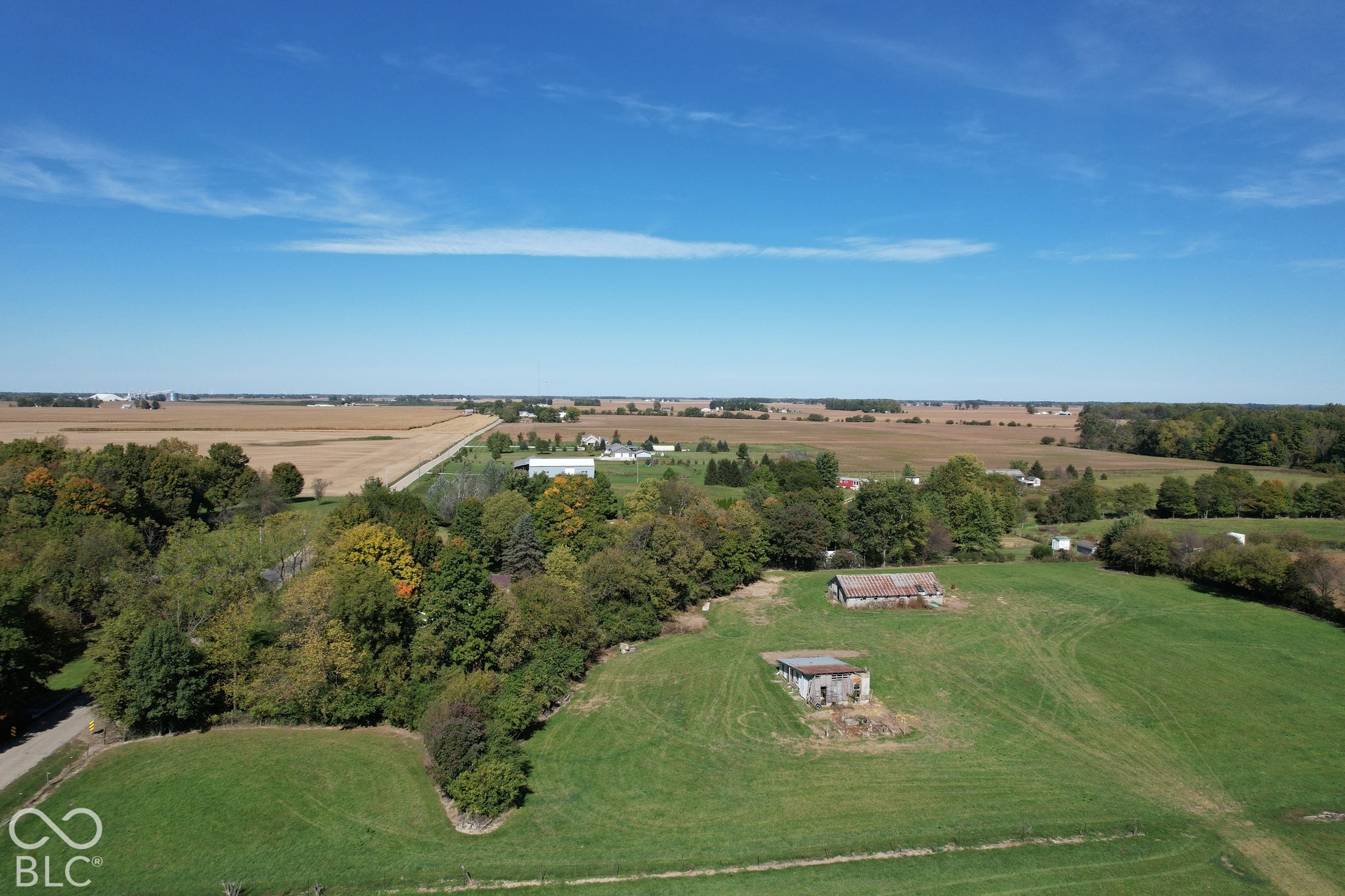 Expansive green fields with two structures, surrounded by mature trees and distant agricultural fields