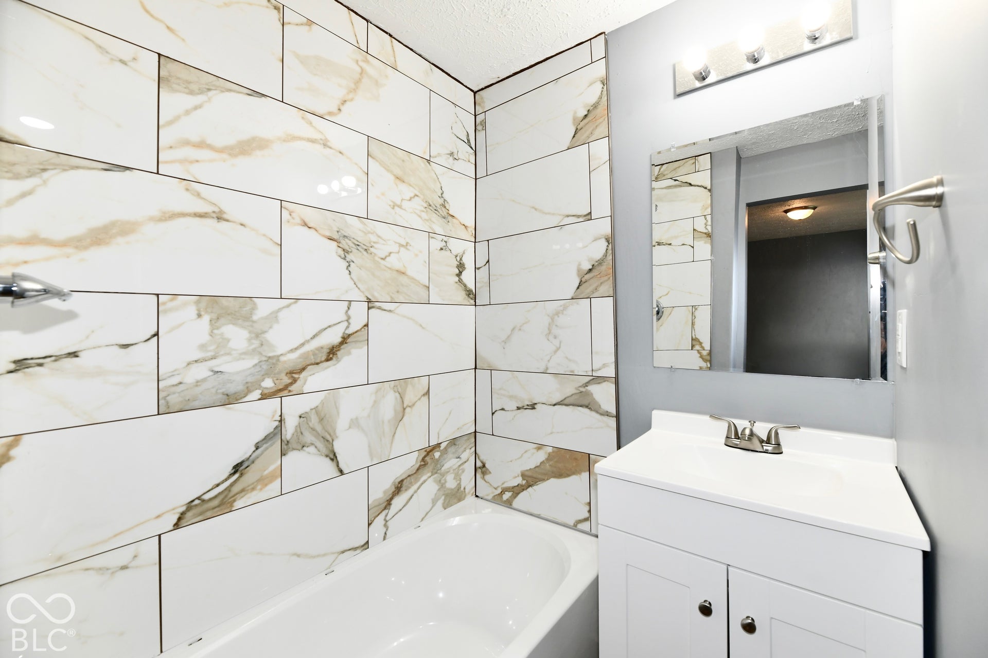 Bathroom featuring large-format decorative tile tub surround, white vanity cabinet with integrated basin, polished metal hardware, vanity mirror, and three-light fixture
