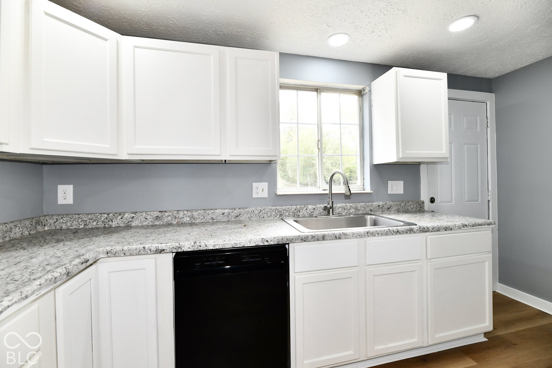Kitchen featuring white shaker-style cabinetry, speckled stone-finish countertops, stainless steel undermount sink with gooseneck faucet, black dishwasher, and wood-finish flooring