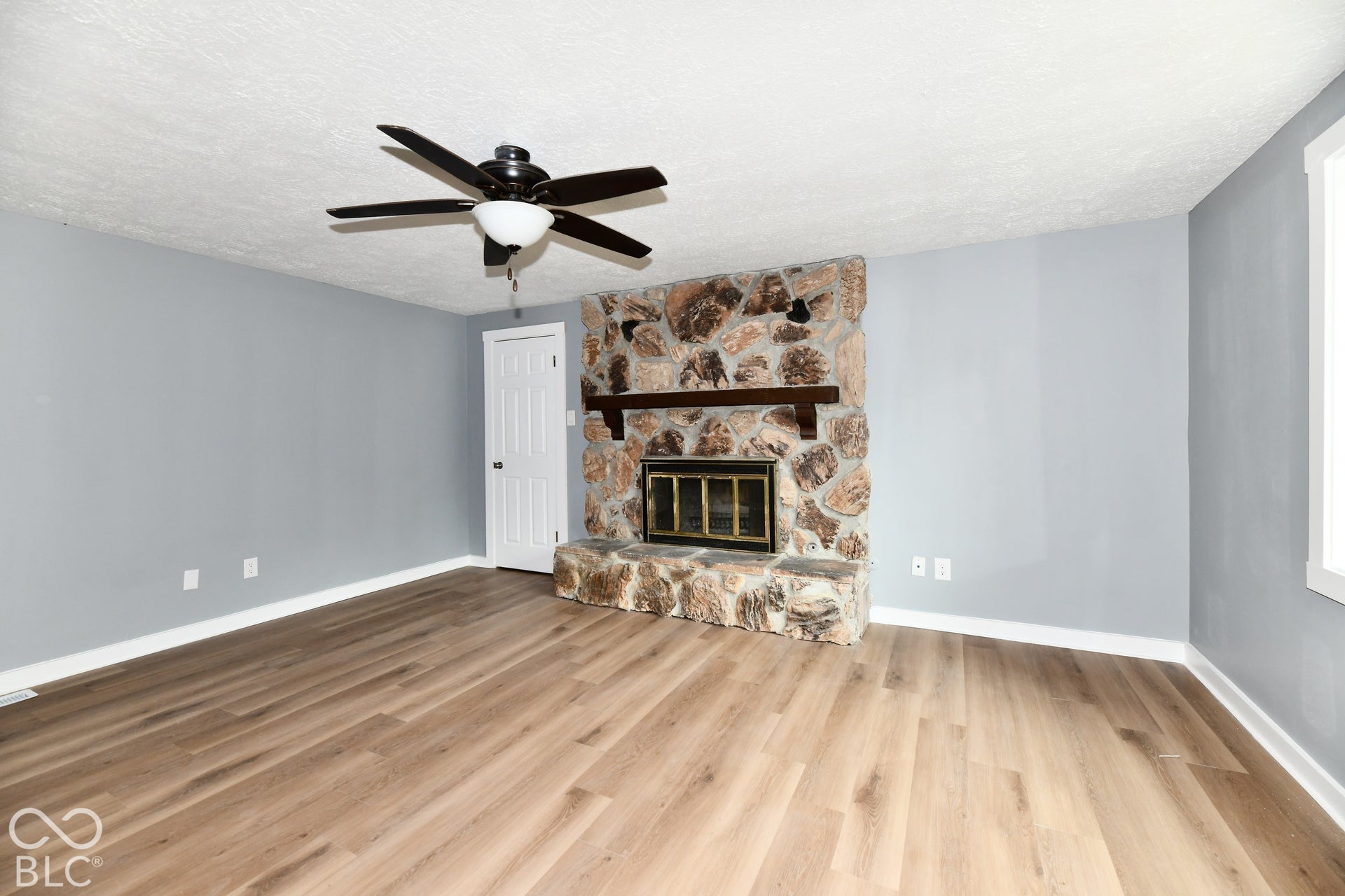 Floor-to-ceiling stone fireplace with a wood mantel, wood-finish flooring, and a ceiling fan with integrated lighting