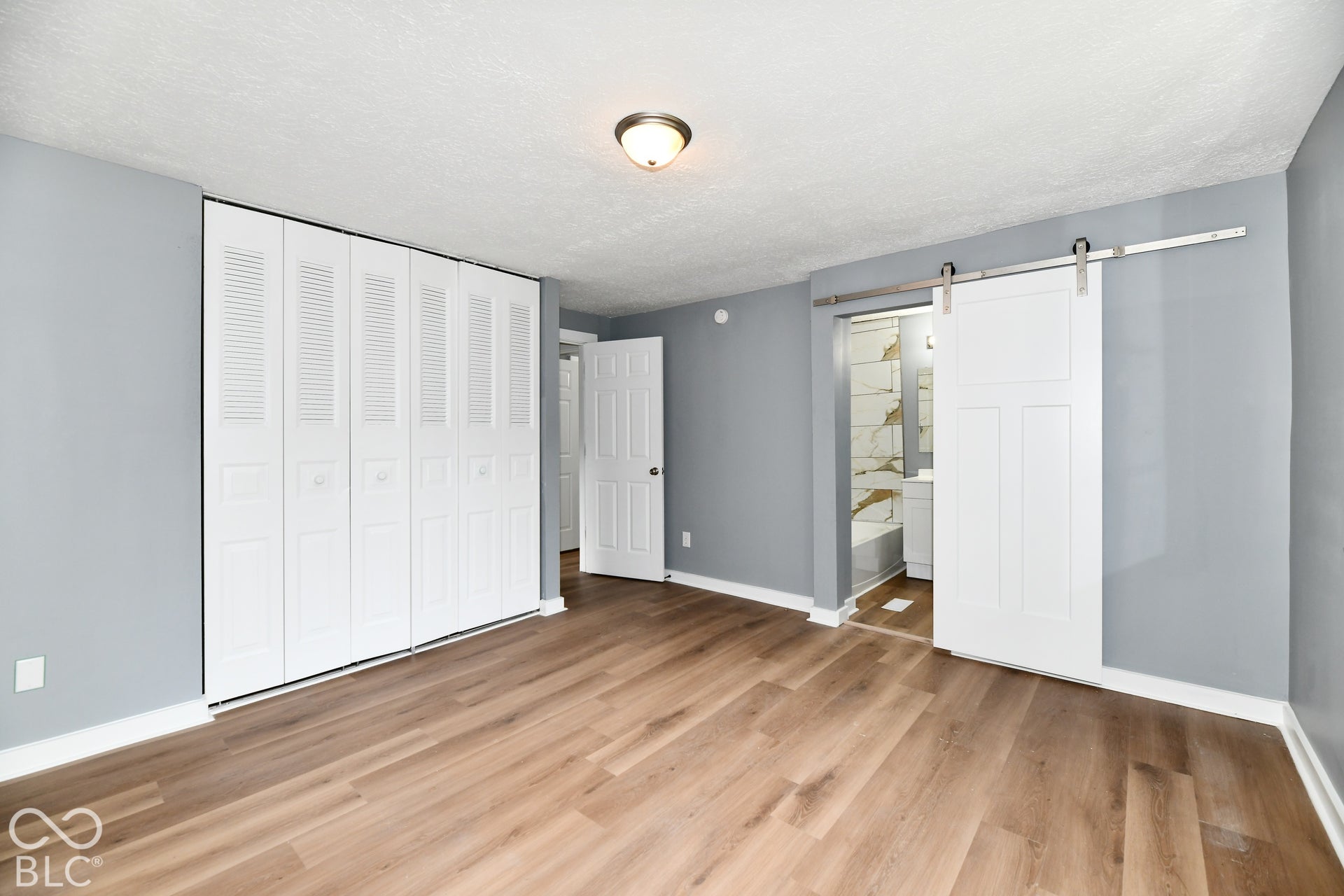 Wood-finish flooring, white louvered bifold closet doors, sliding barn door with stainless steel hardware, and flush-mount ceiling lighting