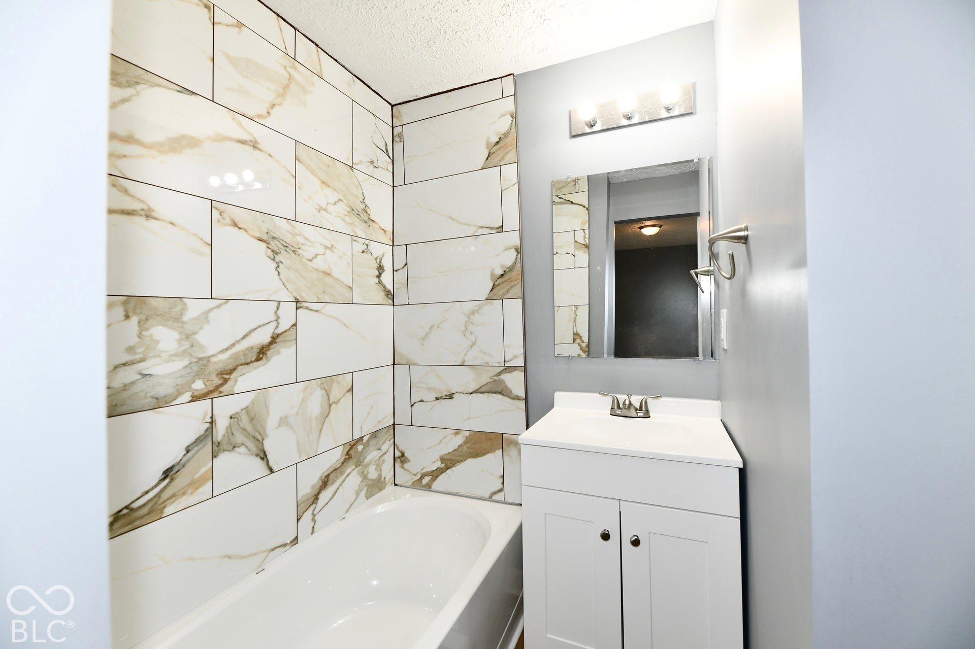 Full-height shower surround featuring large-format veined tile, white vanity cabinet with integrated basin, polished chrome faucet, rectangular wall mirror, and three-light vanity fixture