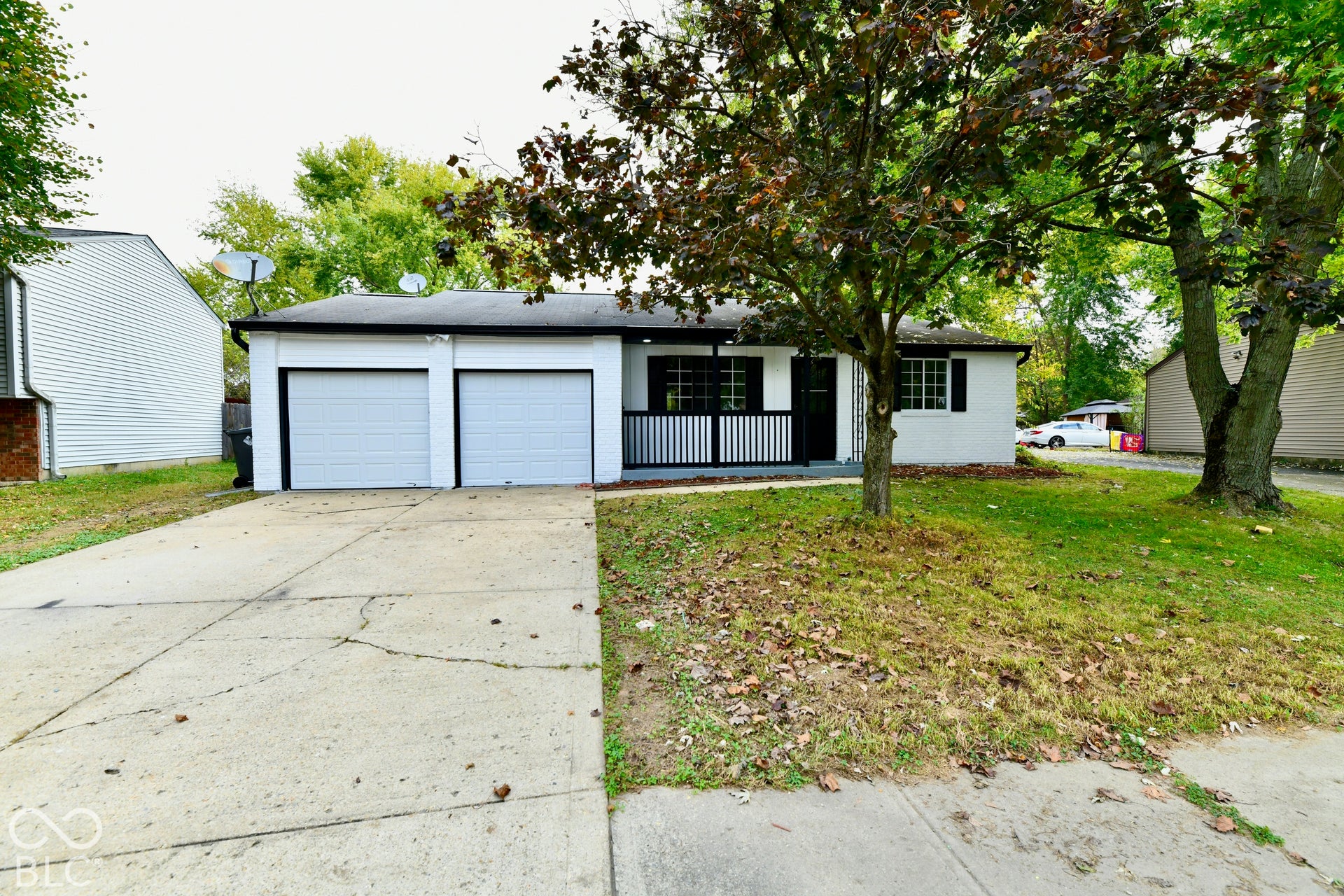 Single-story exterior featuring a two-car attached garage, white painted brick facade, dark-toned shutters, and a covered front porch with iron railing