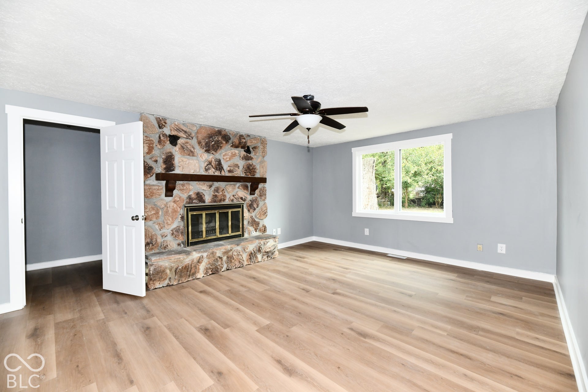 Floor-to-ceiling stone fireplace with wood mantel, wood-finish flooring, and ceiling fan with integrated lighting