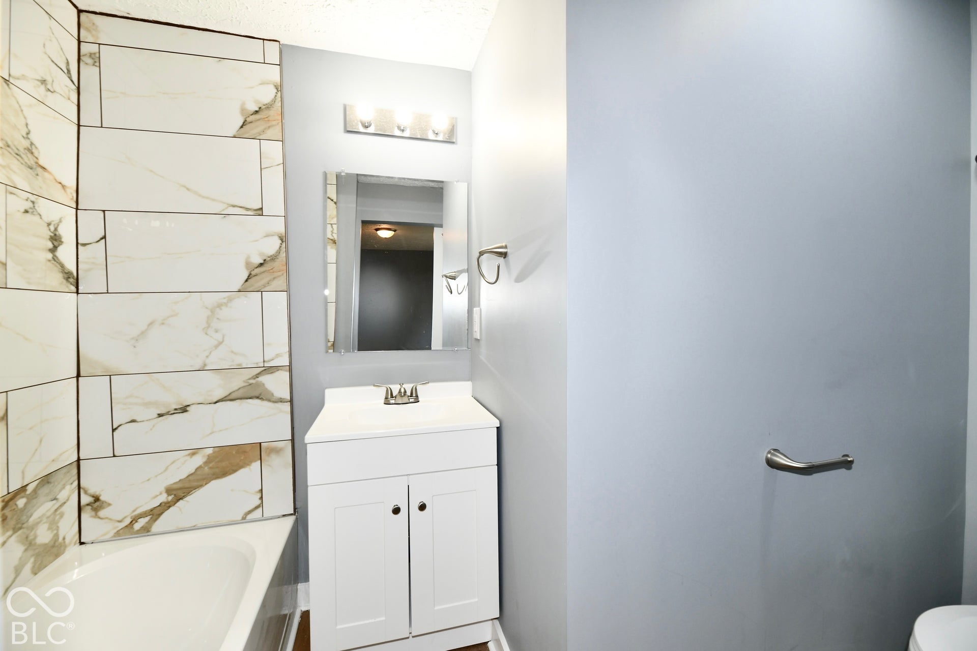 Bathroom featuring white vanity cabinetry, stone-patterned tile wall surround, brushed nickel lighting fixture, and a frameless mirror