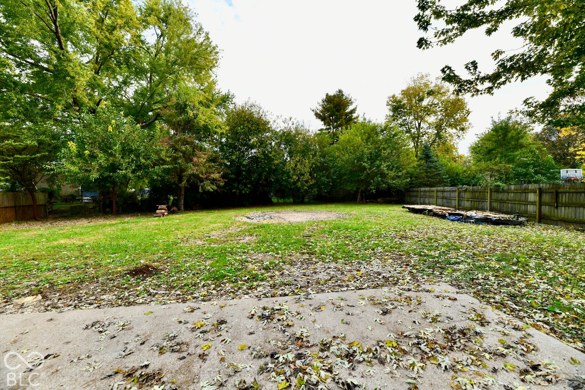 Expansive backyard featuring a concrete patio, a stone-surrounded fire pit, and mature perimeter trees enclosed by wood fencing