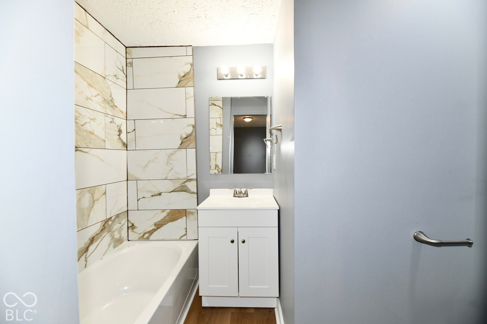 Bathroom featuring a white vanity cabinet with a stone-finish countertop, full-height decorative tile surround, bathtub, wall-mounted mirror, and metal-finish light fixture