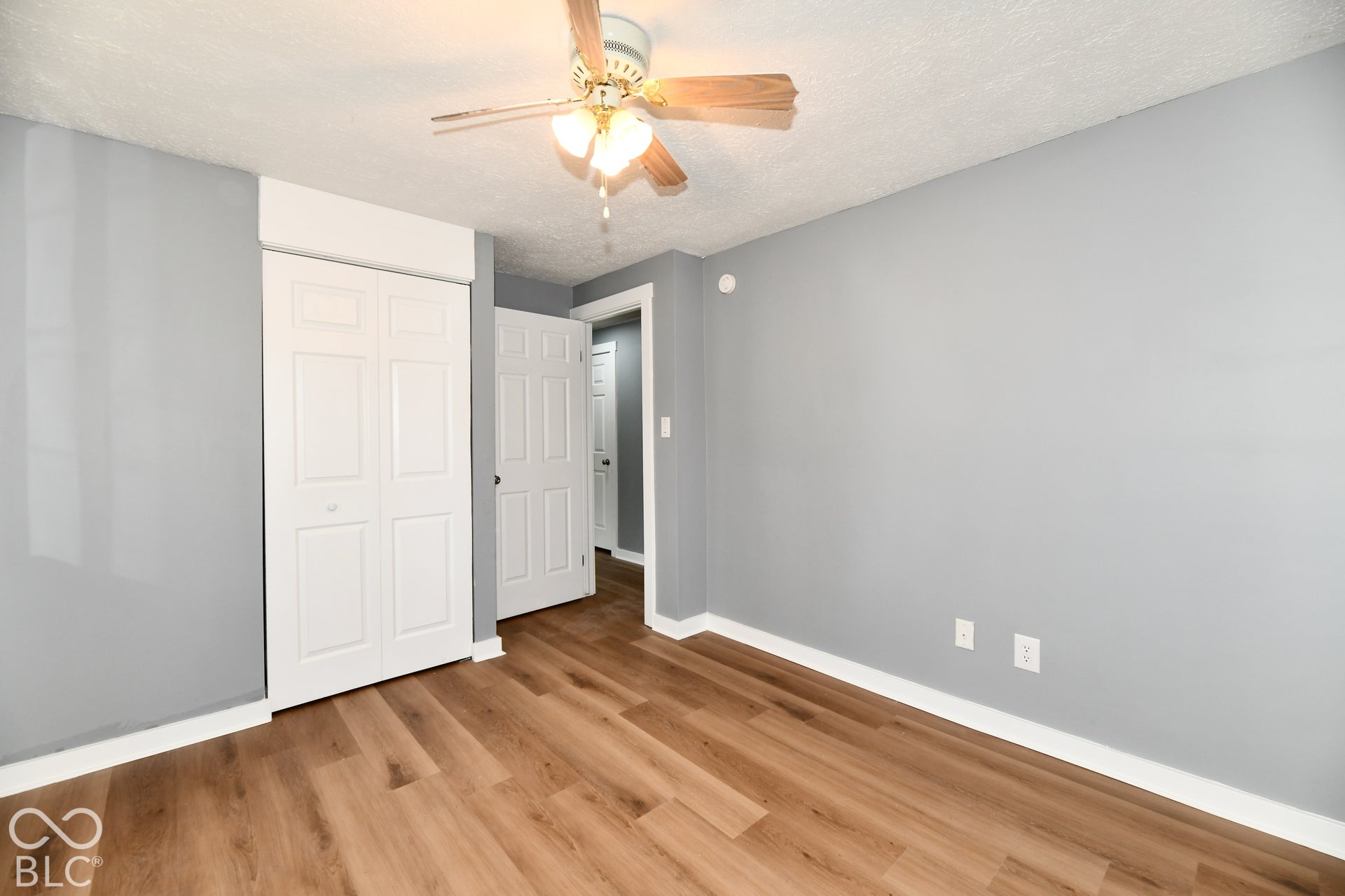 Room featuring wood-finish flooring, neutral gray wall paint, white multi-panel doors, and a ceiling fan with integrated lighting