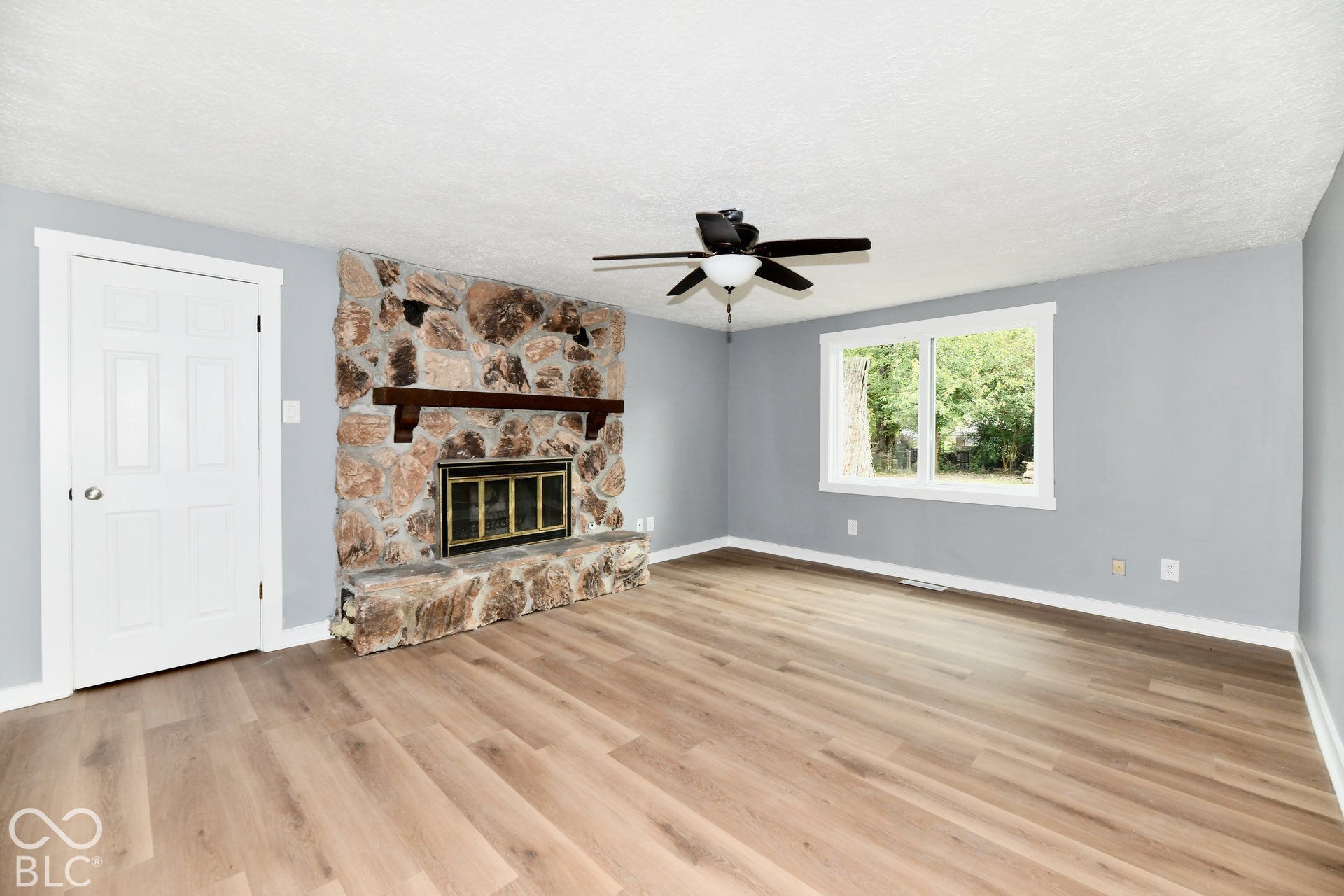 Room featuring wood-finish flooring, a floor-to-ceiling natural stone fireplace with a wood mantel, a multi-pane window, and a ceiling fan