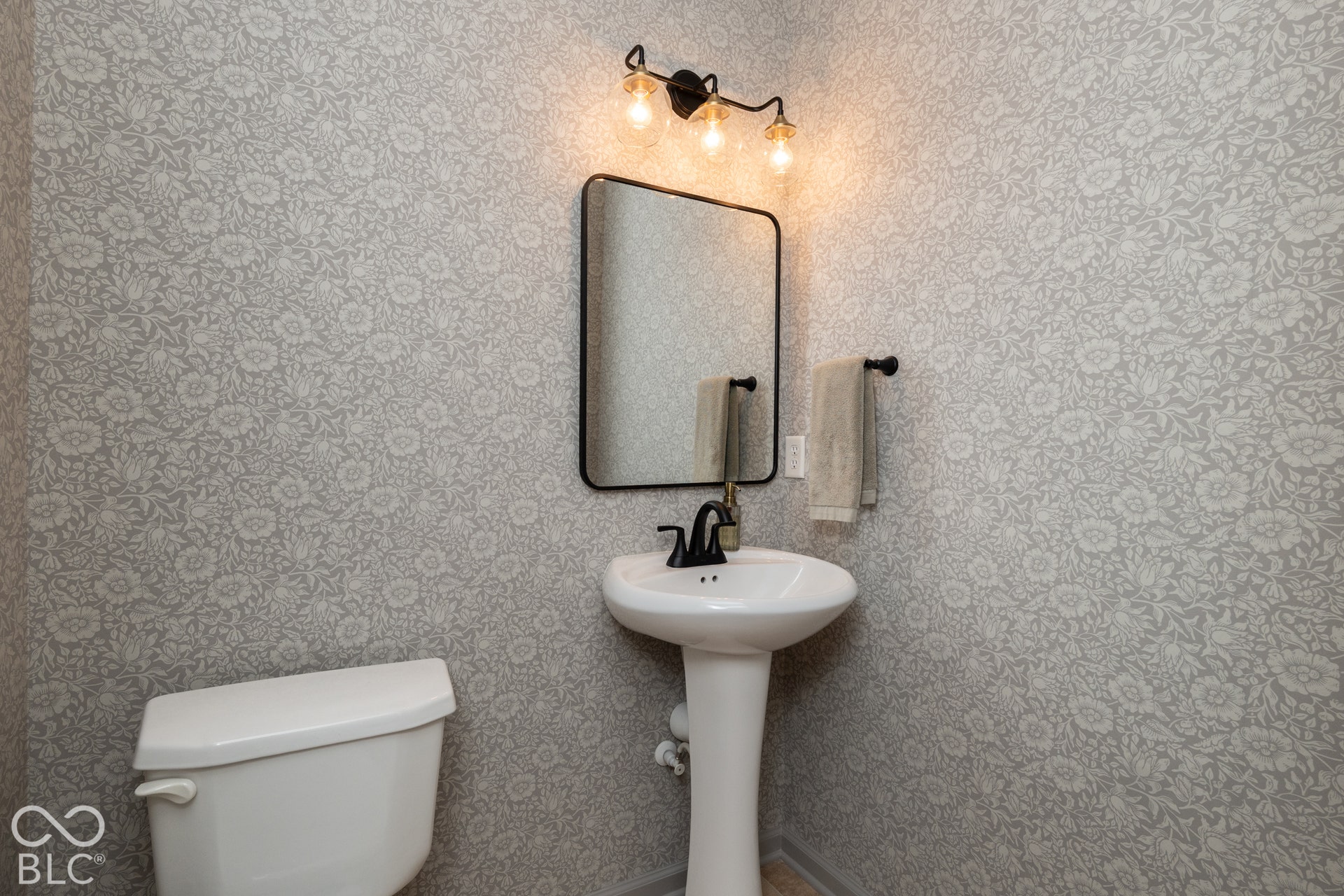 Pedestal sink with a matte black faucet, wall-mounted mirror with a black frame, and a three-light vanity fixture
