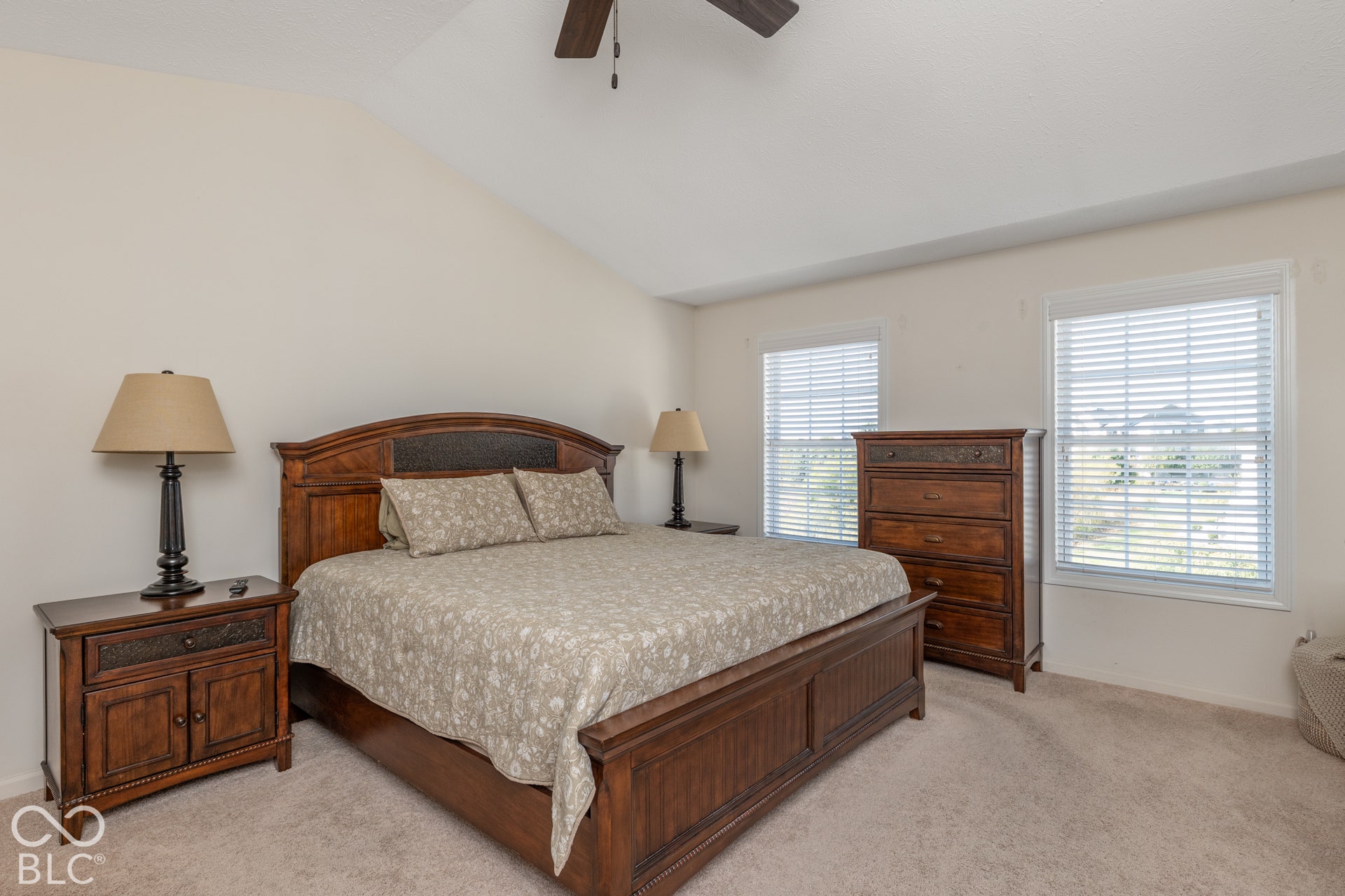 Vaulted ceiling, ceiling fan, two windows with blinds, and neutral carpeting
