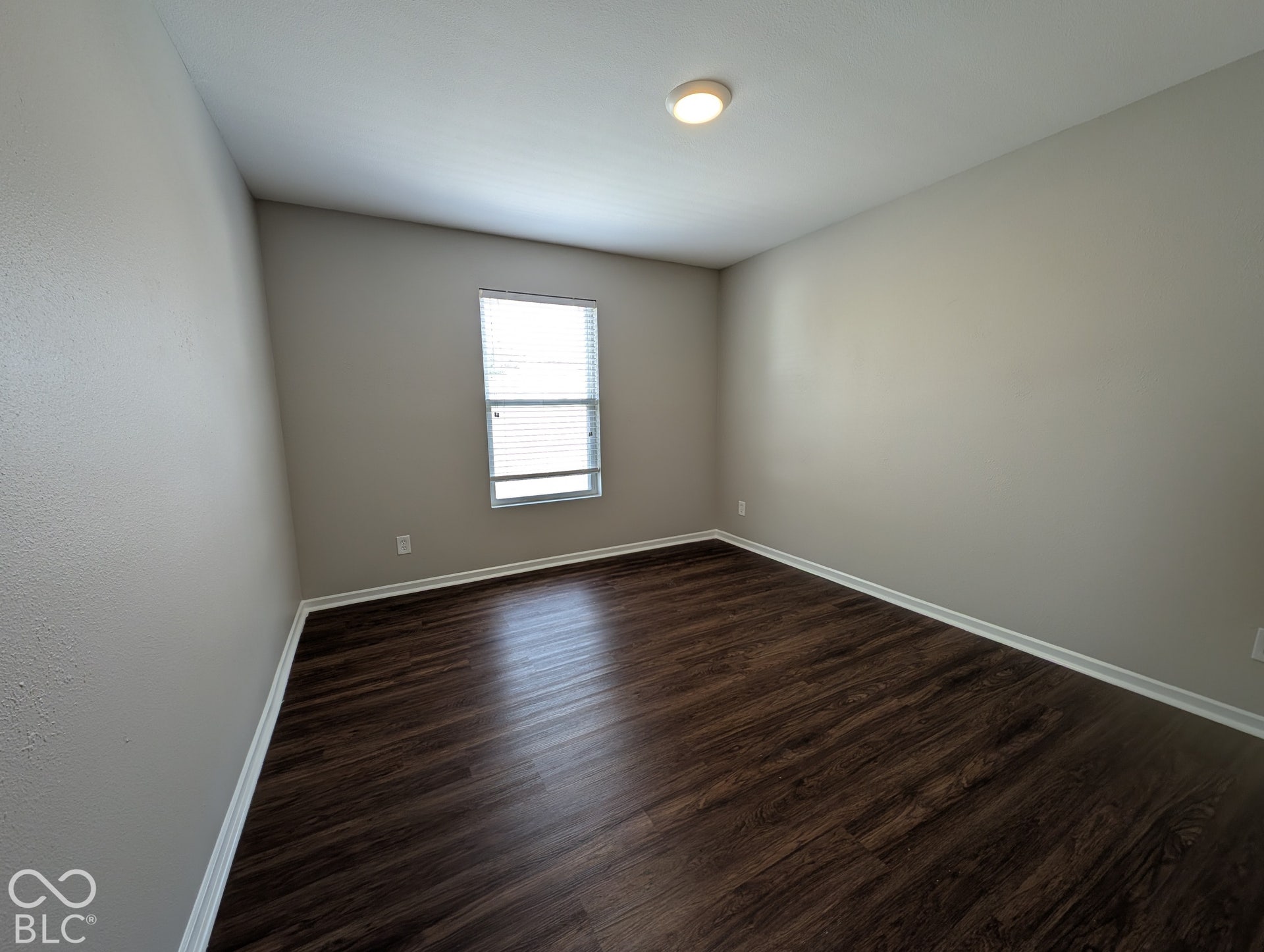 Wood-finish flooring with white trim, a single window with blinds, and a flush-mount ceiling light