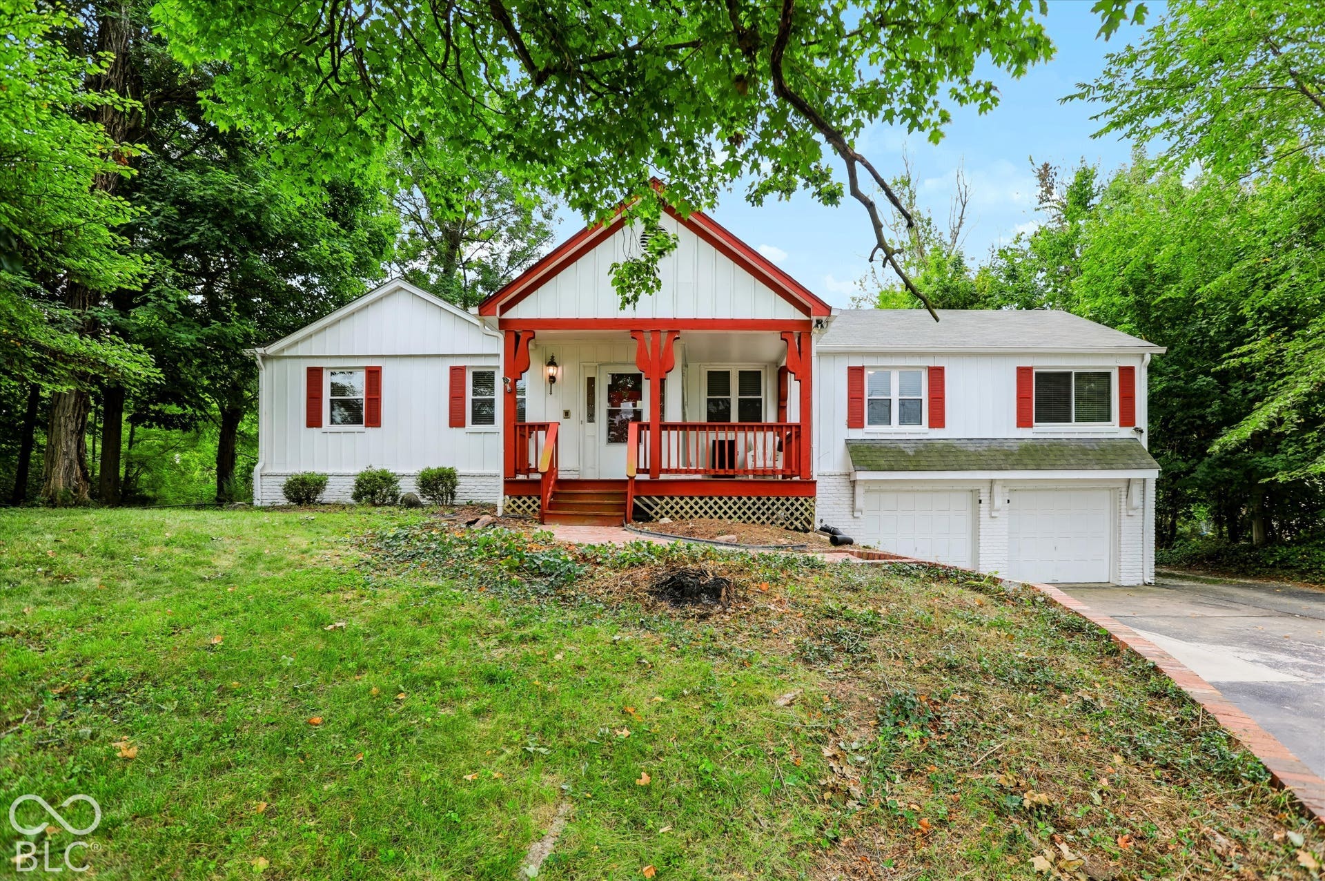 White facade with red trim accents, covered front porch, attached two-car garage, and established perimeter trees
