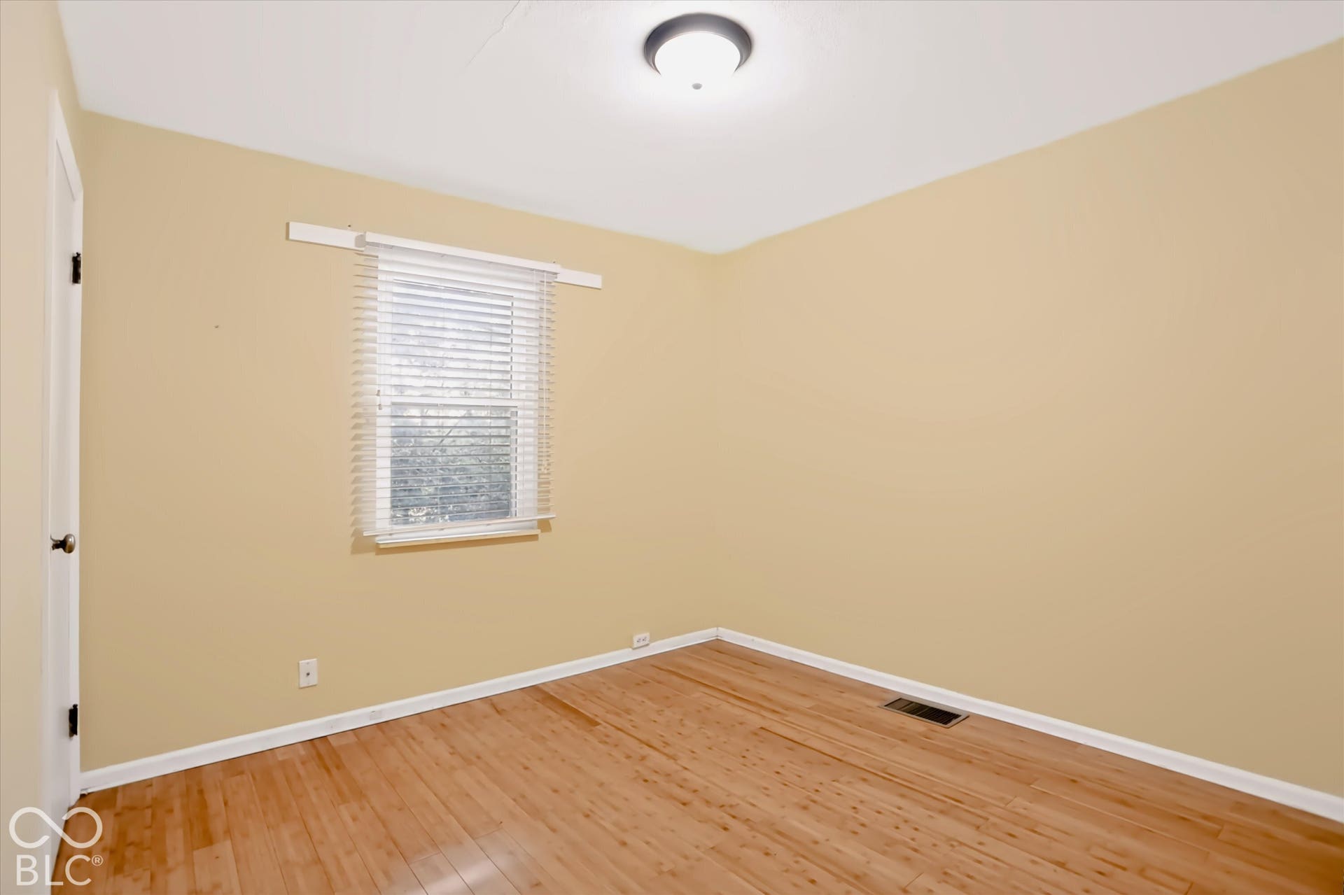 Wood-finish flooring, neutral wall tones, window with horizontal blinds, overhead flush mount light, and white trim