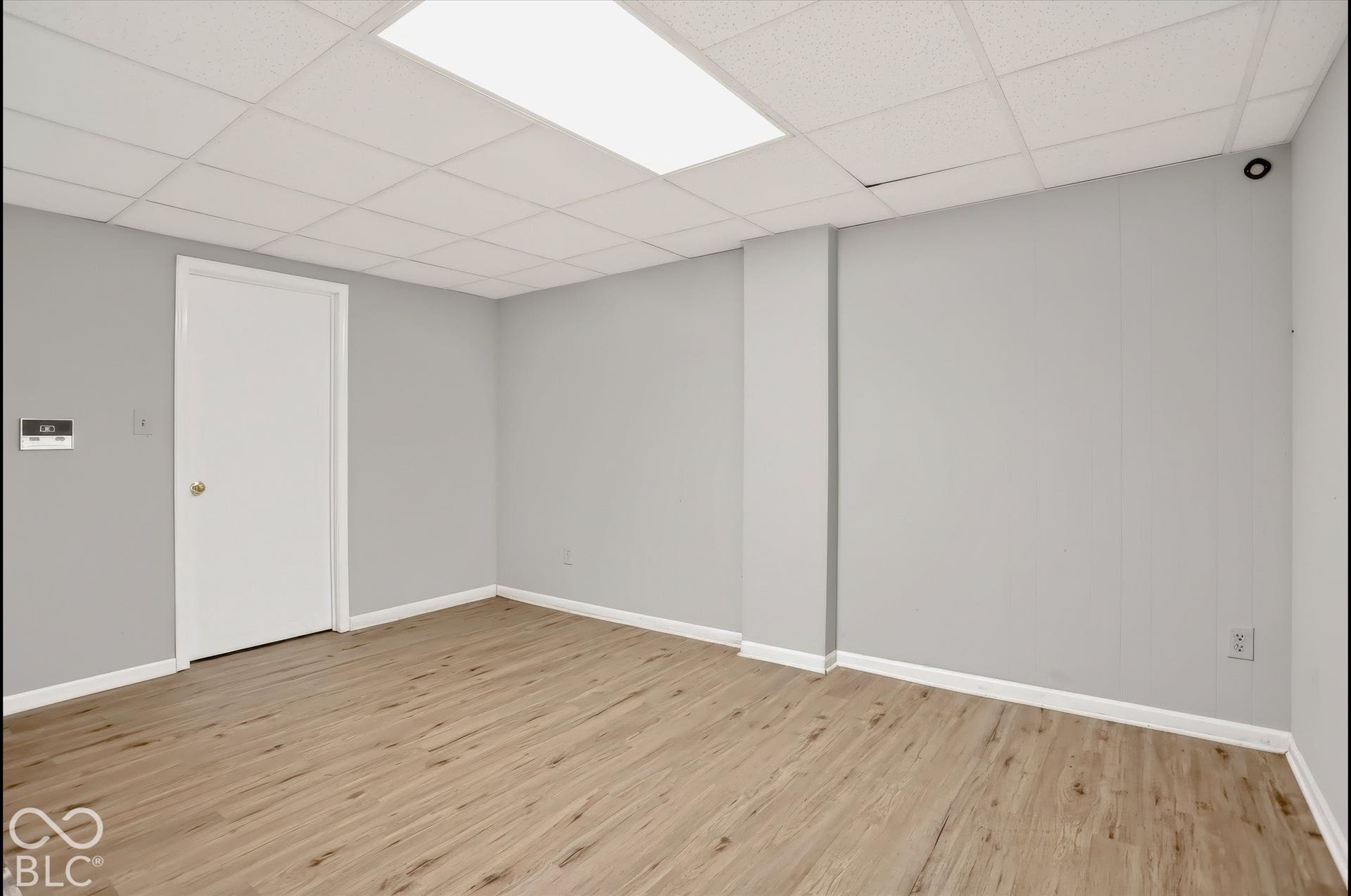 Wood-finish flooring with white trim, light gray walls, and drop ceiling with integrated fluorescent lighting