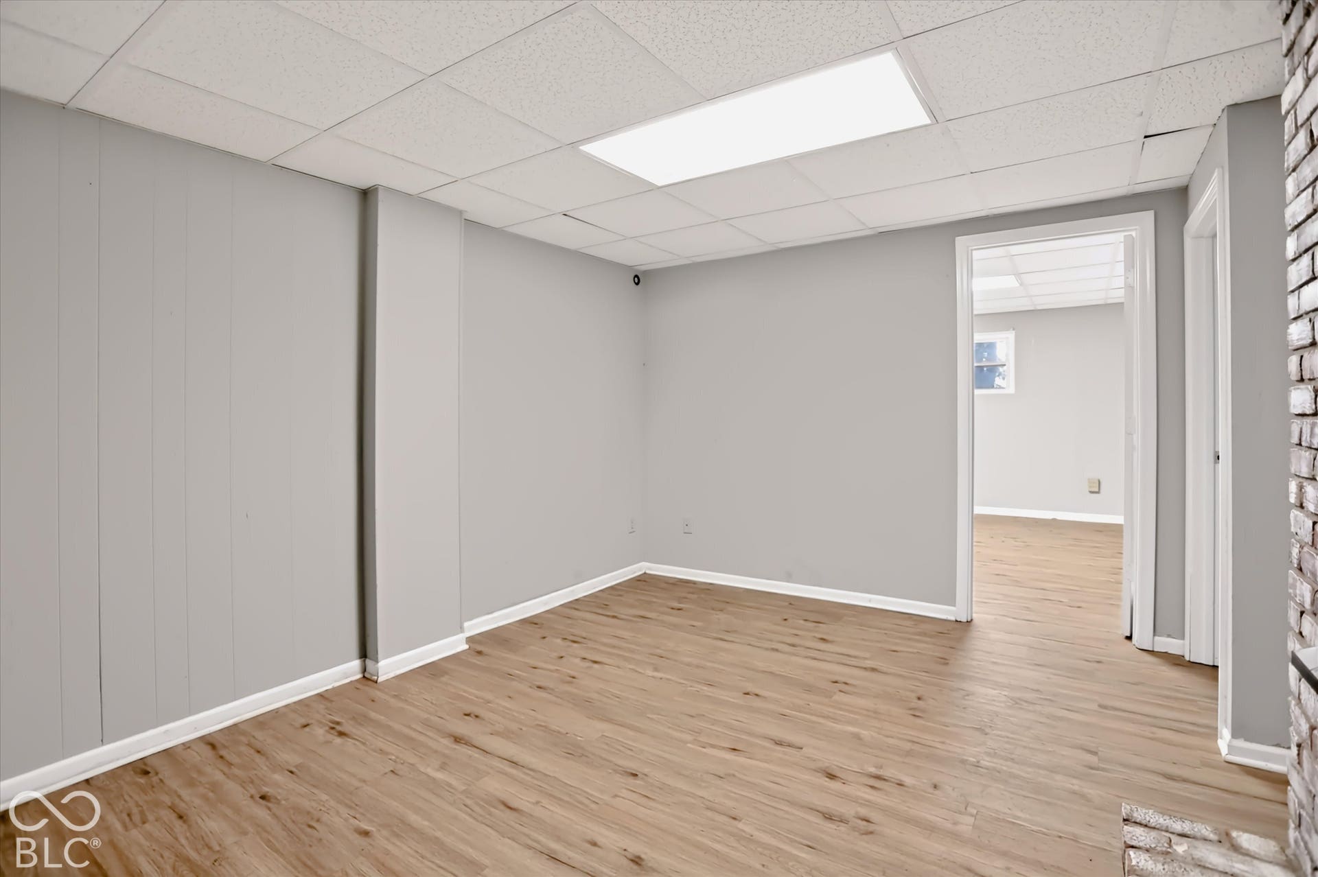Wood-finish flooring, drop ceiling with recessed lighting, light gray wall paneling, and a brick-finish structural element