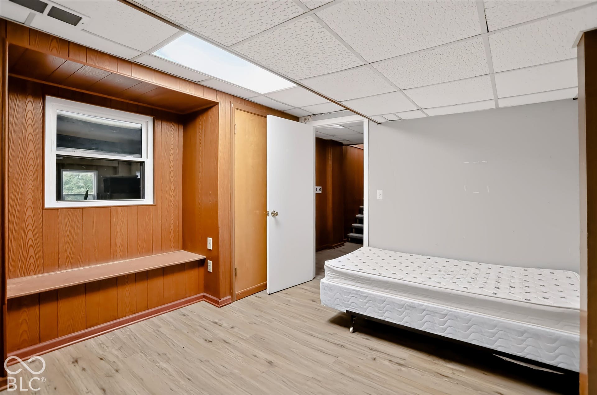 Wood-finish flooring, built-in wooden bench, white-framed window, neutral painted walls, and drop ceiling with recessed lighting