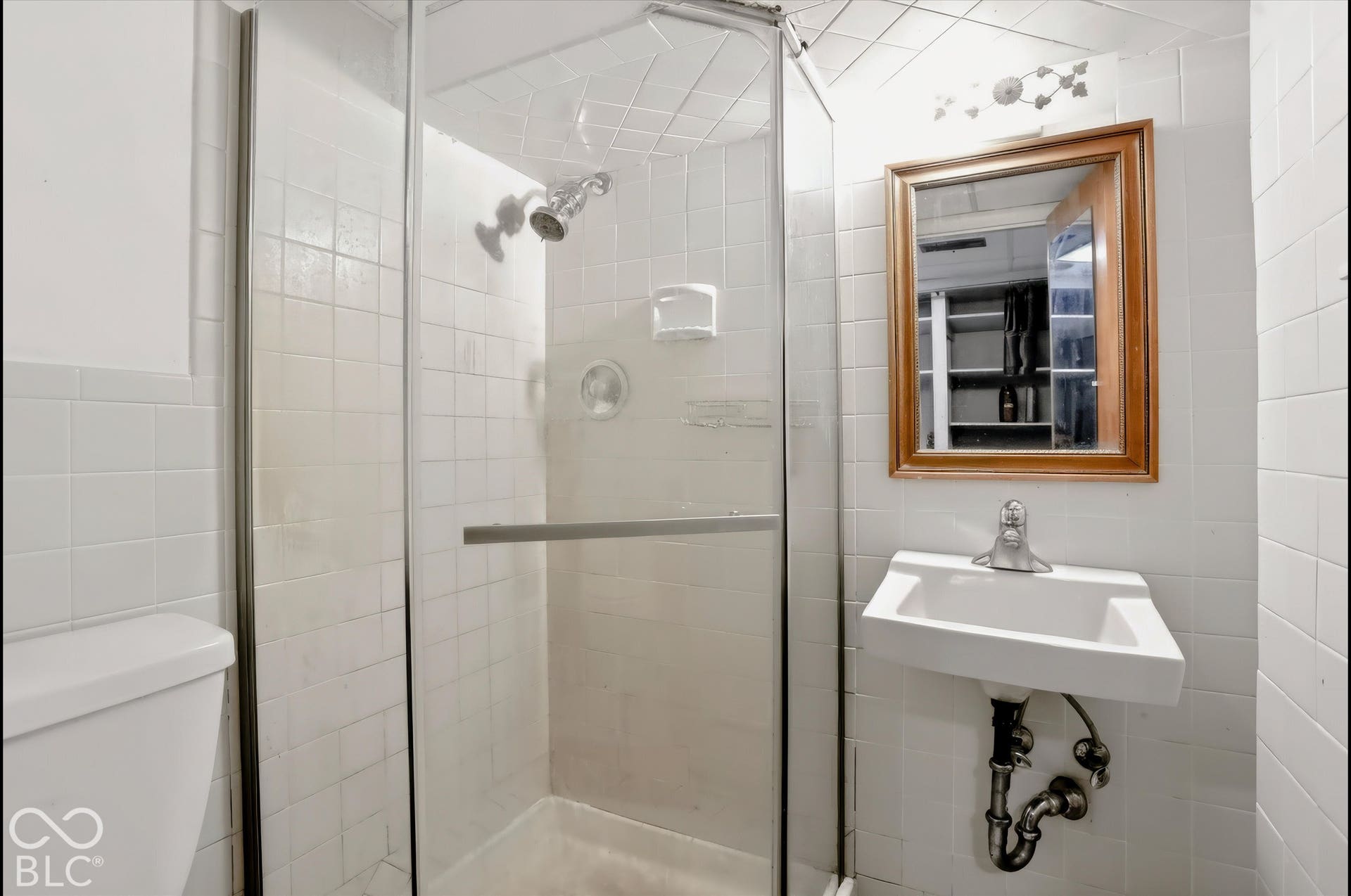 Walk-in shower with clear glass enclosure, wall-mounted shower head, white tiled walls, wall-mounted sink with chrome faucet, and framed mirror