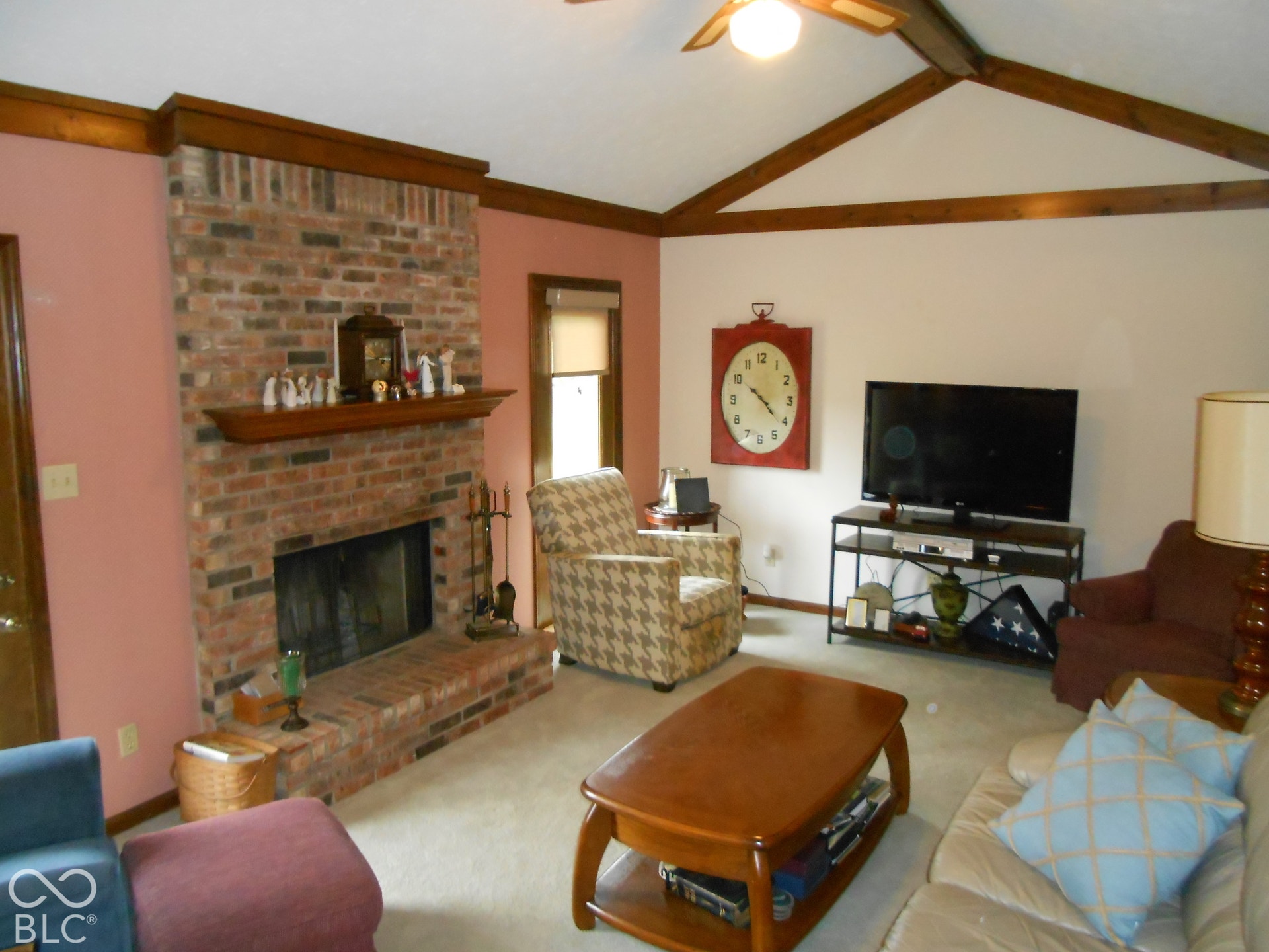 Brick fireplace with a wood mantle, vaulted ceiling with wood beams, and a ceiling fan