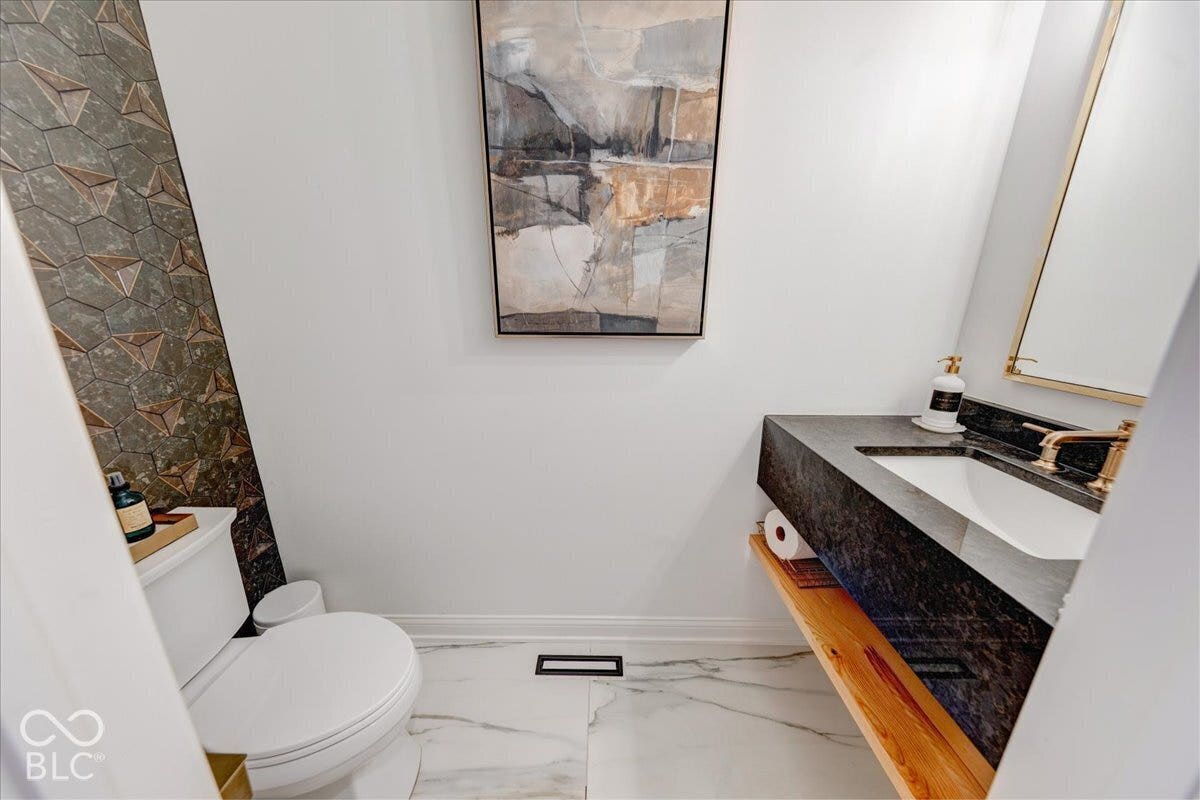 White porcelain toilet, marble-look floor tile, textured wall tile, wall-mounted sink with dark stone countertop, and brass-finish faucet