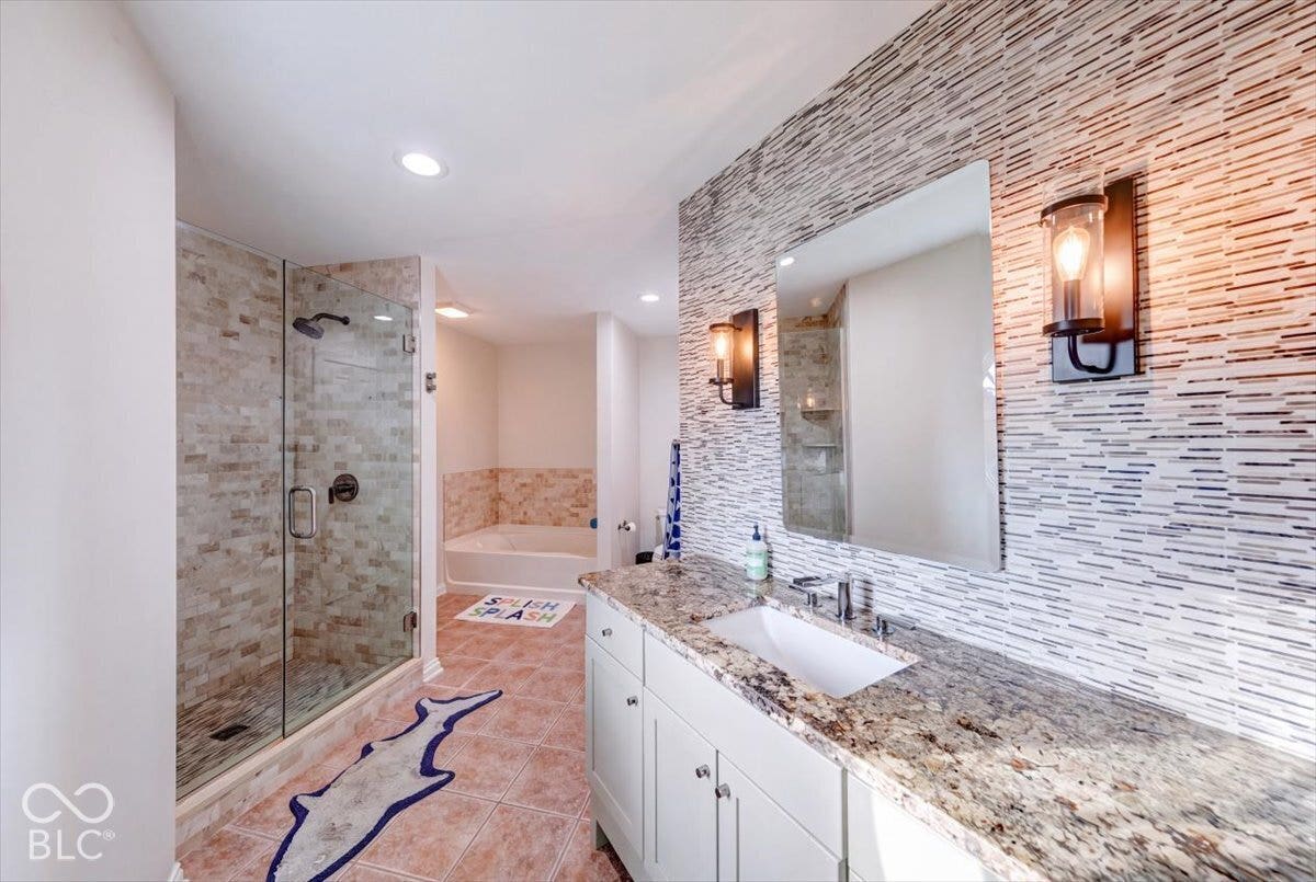 Spa-inspired primary bathroom featuring a walk-in shower with linear tile surround and frameless glass enclosure, dual-sink vanity with white cabinetry and granite countertops, linear tile accent wall with integrated sconce lighting, and recessed ceiling lights throughout