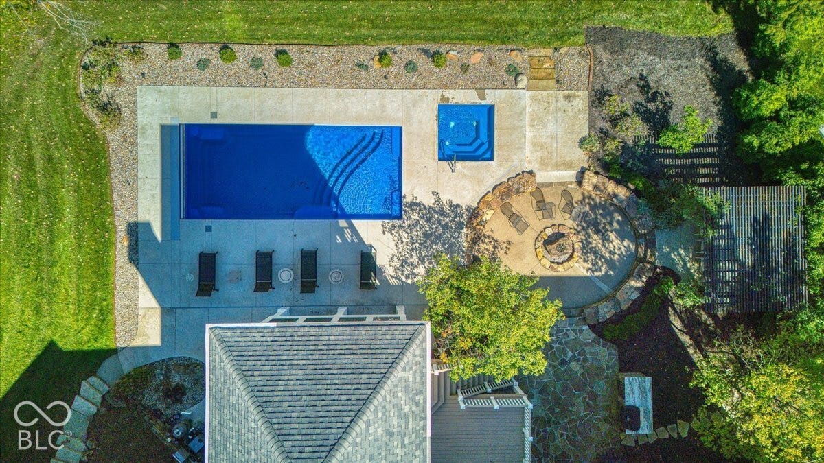 In-ground swimming pool with an integrated spa, extensive concrete patio, stone-paved fire pit area, and mature landscaping