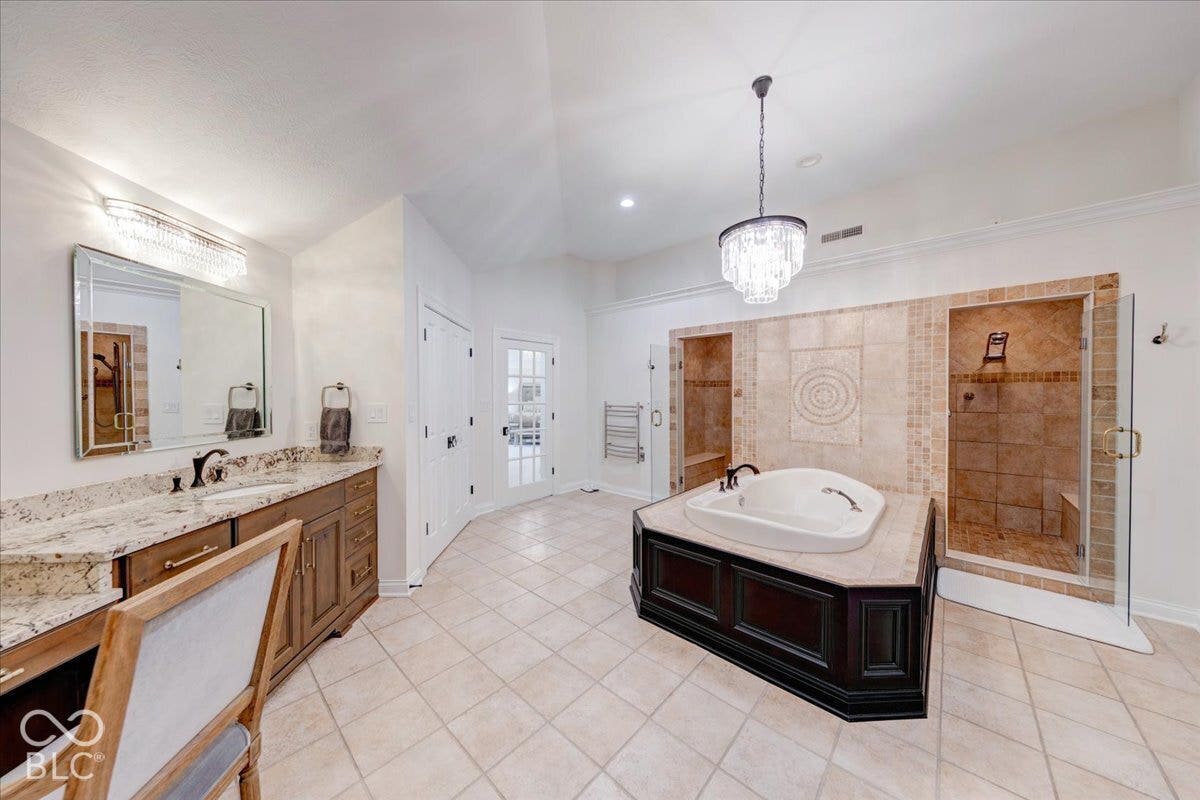 Dual walk-in showers with glass enclosures, a central soaking tub, tile flooring, a dual vanity with a granite countertop, and a crystal chandelier