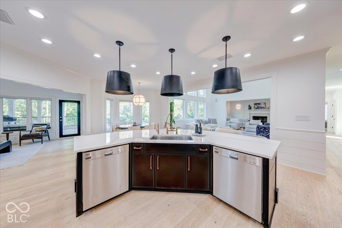 Kitchen island featuring a stainless steel sink, dual built-in dishwashers, dark cabinetry with copper hardware, and a light-toned countertop
