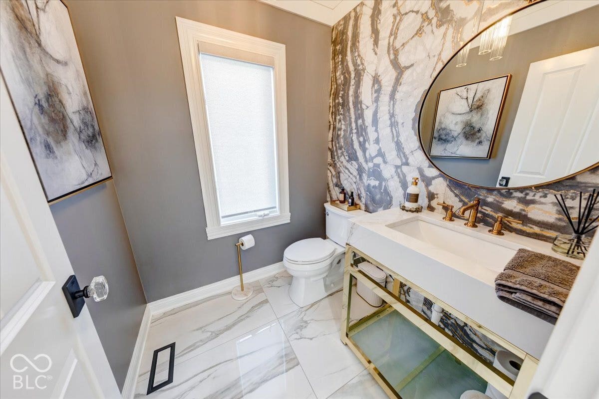 Powder room featuring a large round mirror, a vanity with a white countertop and gold-tone base, and a wall with a distinctive stone-look finish