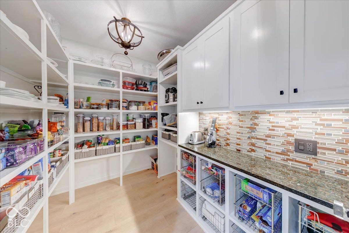 Custom pantry featuring extensive built-in shelving, wood-finish flooring, a decorative ceiling fixture, and a countertop area with mosaic tile backsplash and upper cabinetry