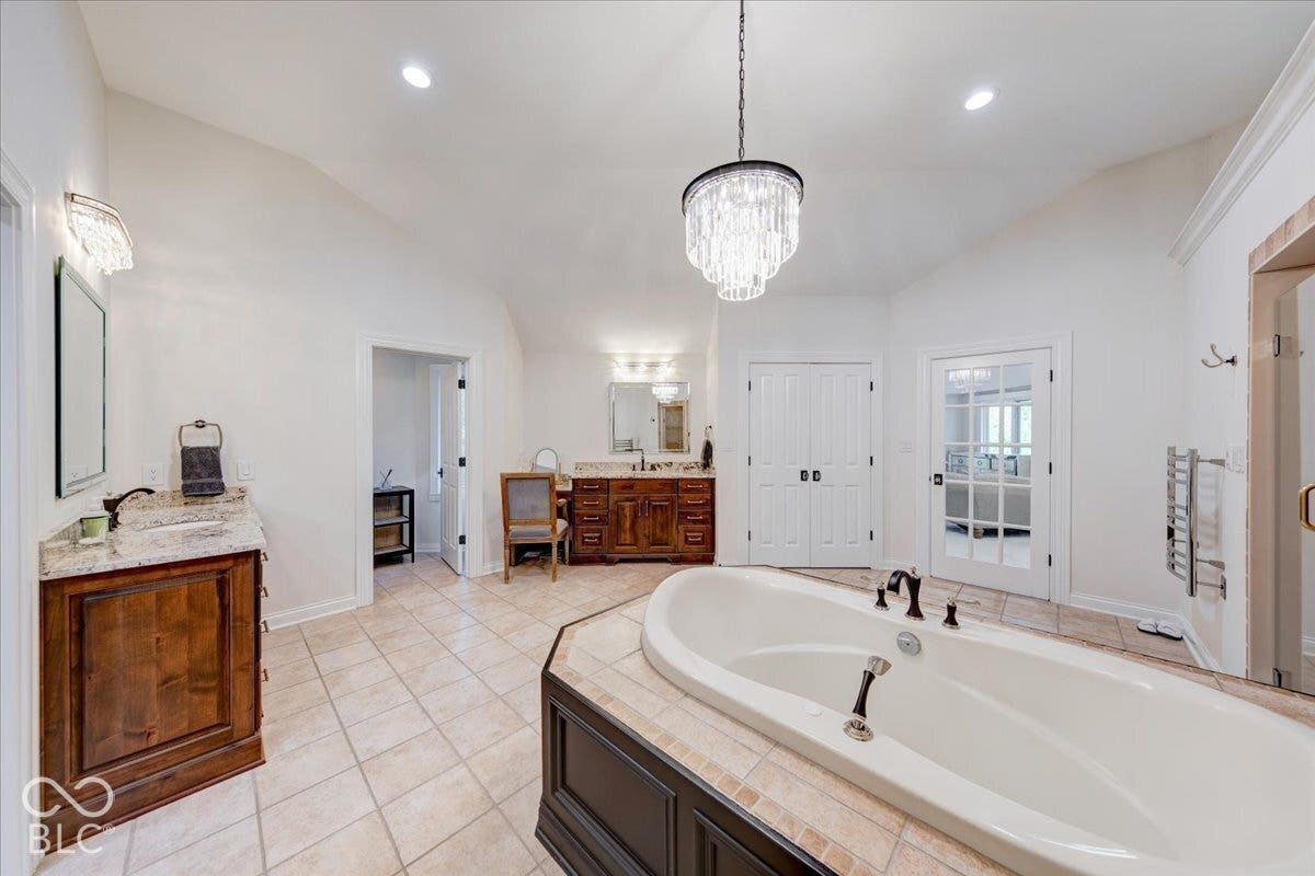 Luxurious primary bathroom featuring a freestanding soaking tub with deck-mounted fixtures, dual vanities with wood cabinetry and stone countertops, crystal chandelier pendant lighting, tile flooring, and multiple entry doors providing convenient access throughout the suite