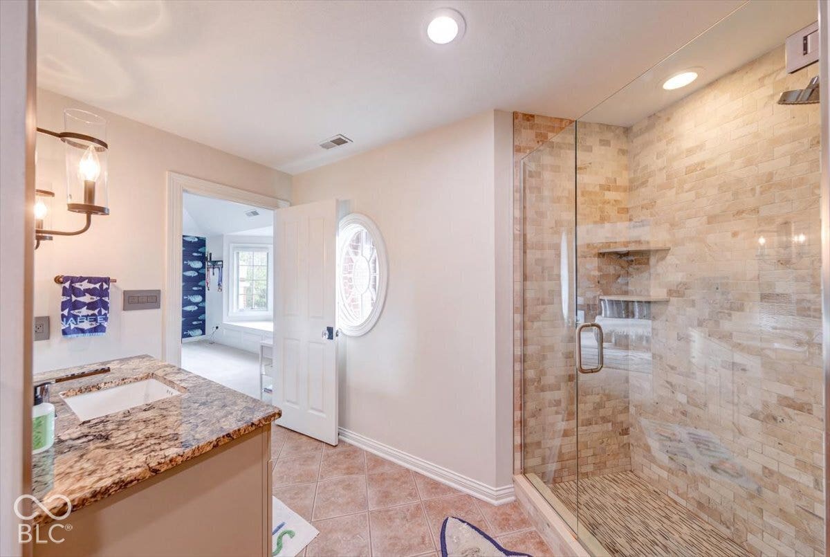 Spa-inspired primary bathroom featuring a luxurious soaker tub with granite surround, dual vanity with stone countertops, and a frameless glass-enclosed walk-in shower with natural stone tile detailing