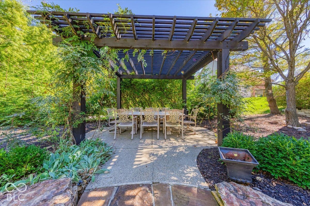 Expansive outdoor living area featuring a large dark-stained wood pergola over a paved patio, surrounded by mature trees and varied landscaping