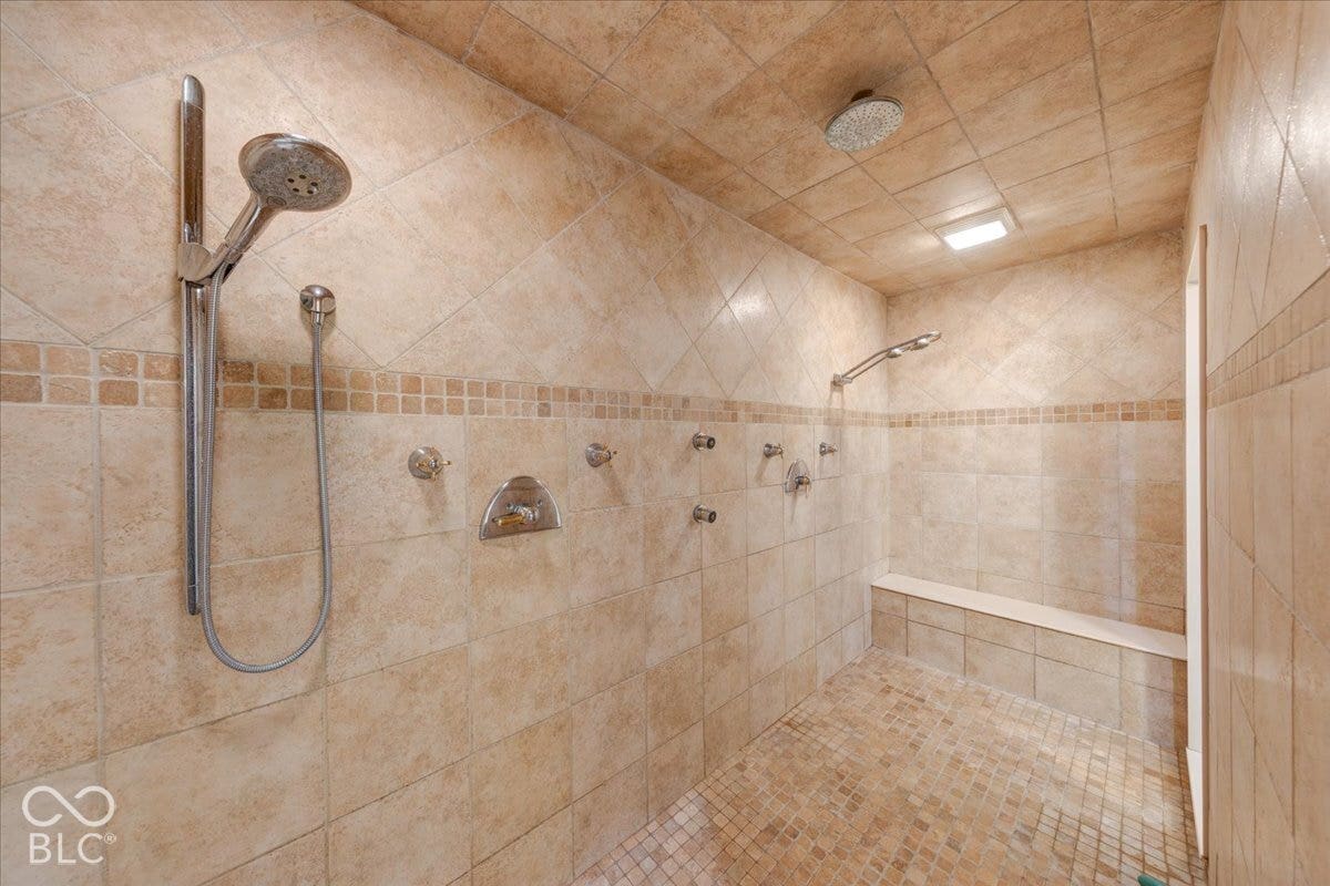 Spa-inspired primary bathroom featuring a spacious walk-in shower with travertine tile surround, dual showerheads, handheld spray fixture, and integrated niche shelving