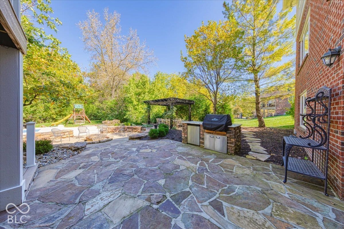 Expansive flagstone patio with an outdoor kitchen featuring built-in grill and stainless steel cabinetry, a stone-lined fire pit area, and a wooden pergola