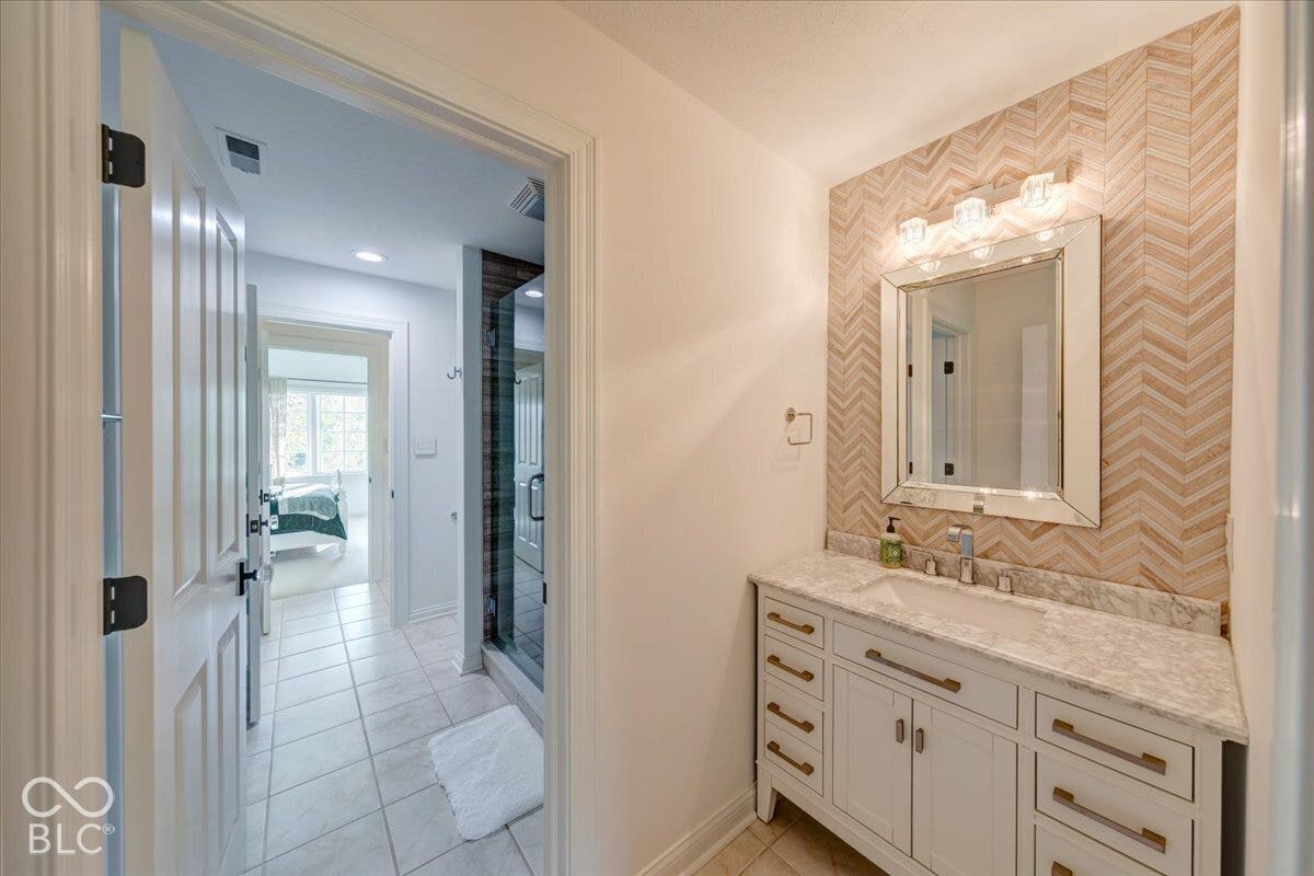 Primary bathroom featuring dual vanity with brushed brass hardware, white cabinetry with ample storage, marble countertops, herringbone tile backsplash, framed mirror with integrated lighting, and an adjoining spa-like ensuite with walk-in shower and white subway tile flooring