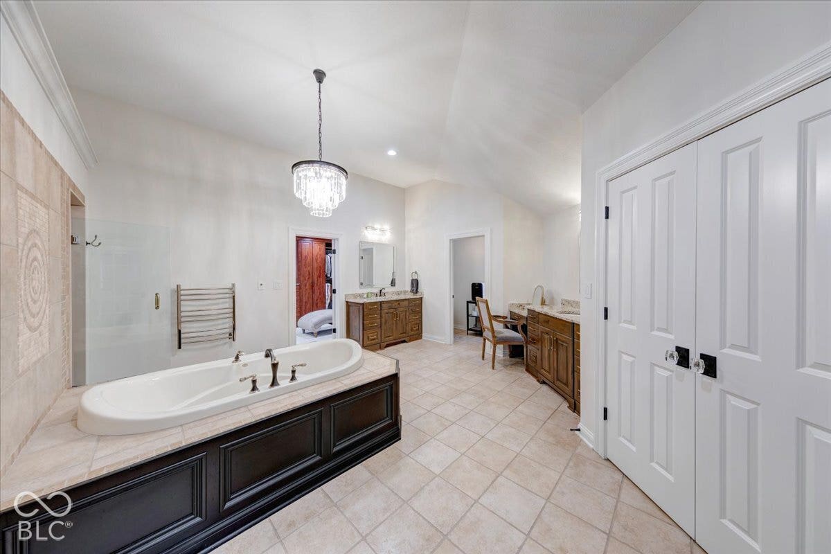 Soaking tub with bronze fixtures, dual wood-finish vanities, elegant chandelier, vaulted ceiling, and panel-style doors