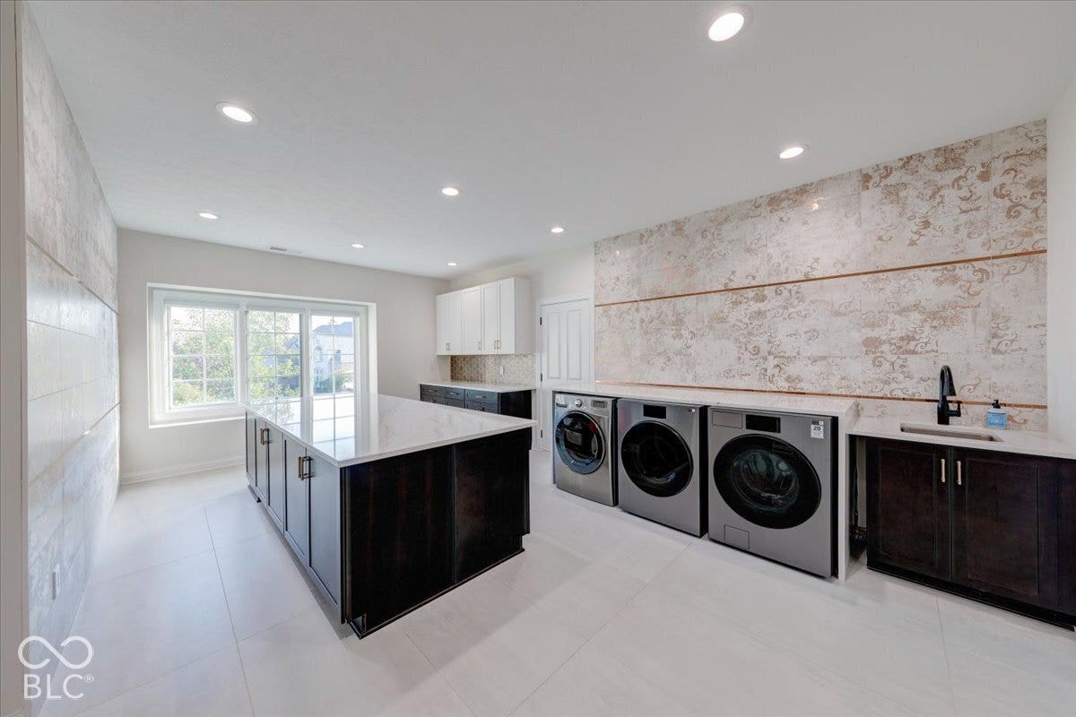 Luxurious laundry room featuring three stainless steel front-load washers and dryers, sleek black cabinetry with integrated sink, and decorative tile backsplash