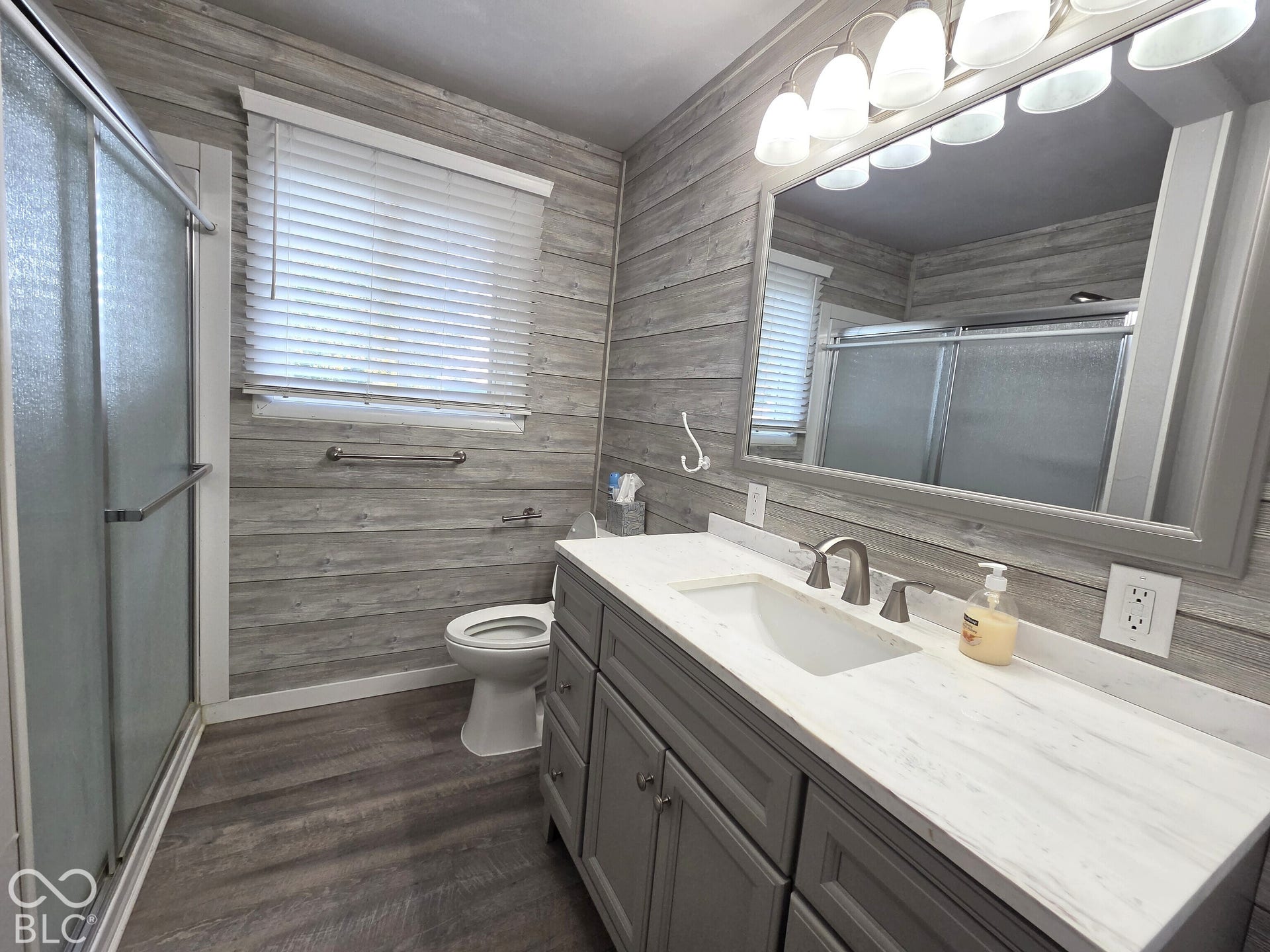1221 Elk Lane, Elwood, IN 46036 Bathroom featuring a grey vanity with a white countertop, an integrated rectangular sink, wood-finish wall paneling, a shower enclosure with sliding glass doors, and a large framed wall mirror
