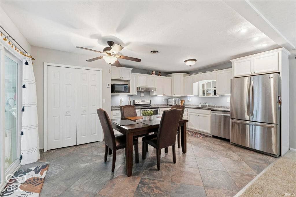 Spacious kitchen and dining area featuring stainless steel appliances, white cabinetry with ample storage, stone-finish flooring throughout, and a ceiling fan with integrated lighting