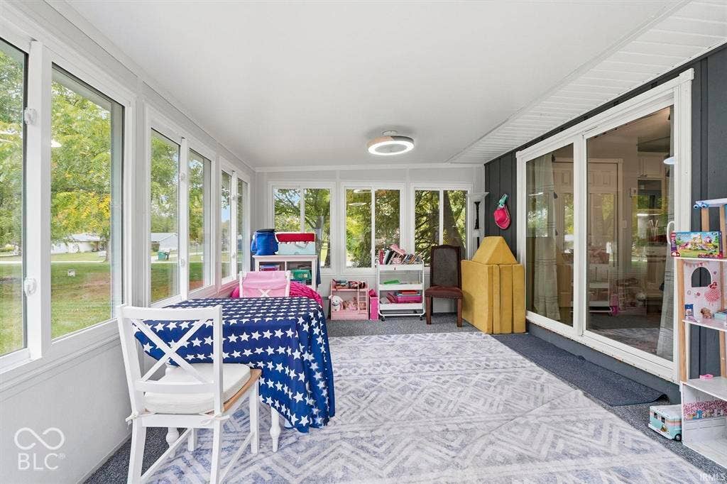 Bright sunroom with expansive multi-panel windows framing natural landscape views, geometric patterned area rug, white ceiling with recessed lighting, and direct access to outdoor spaces through sliding glass doors