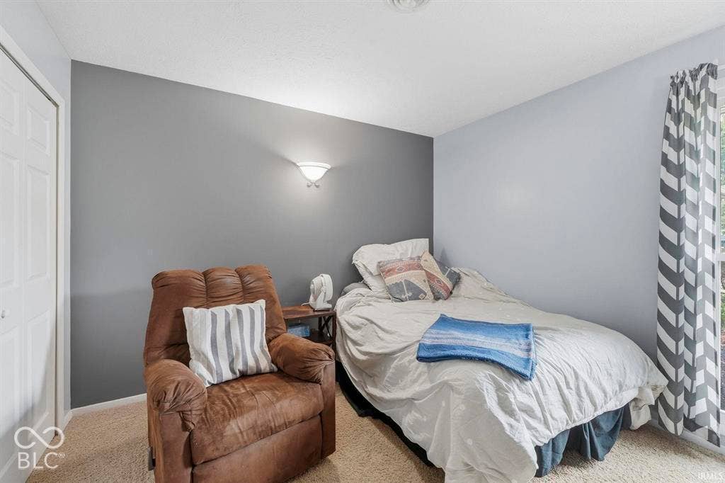 Bedroom featuring neutral wall finishes with an accent gray feature wall, carpeted flooring, and abundant natural light from a corner window with striped roman shade
