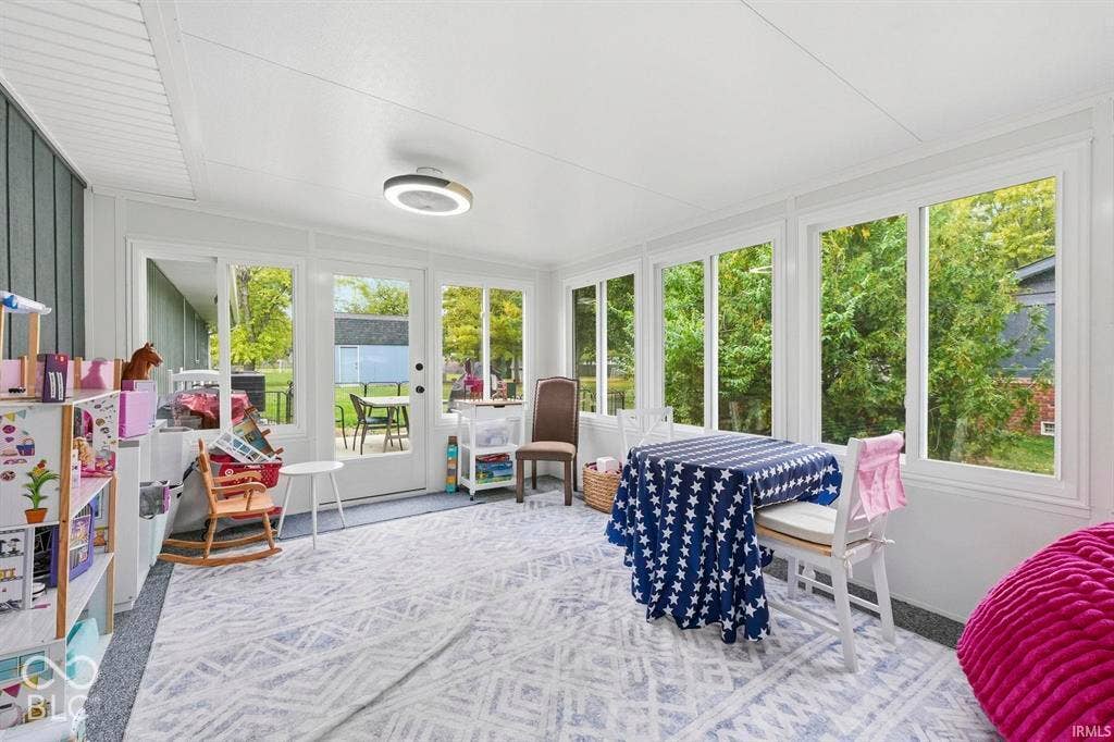 Bright, light-filled sunroom with expansive multi-panel windows framing mature landscaping, geometric patterned flooring, recessed ceiling lighting, and direct access to exterior spaces