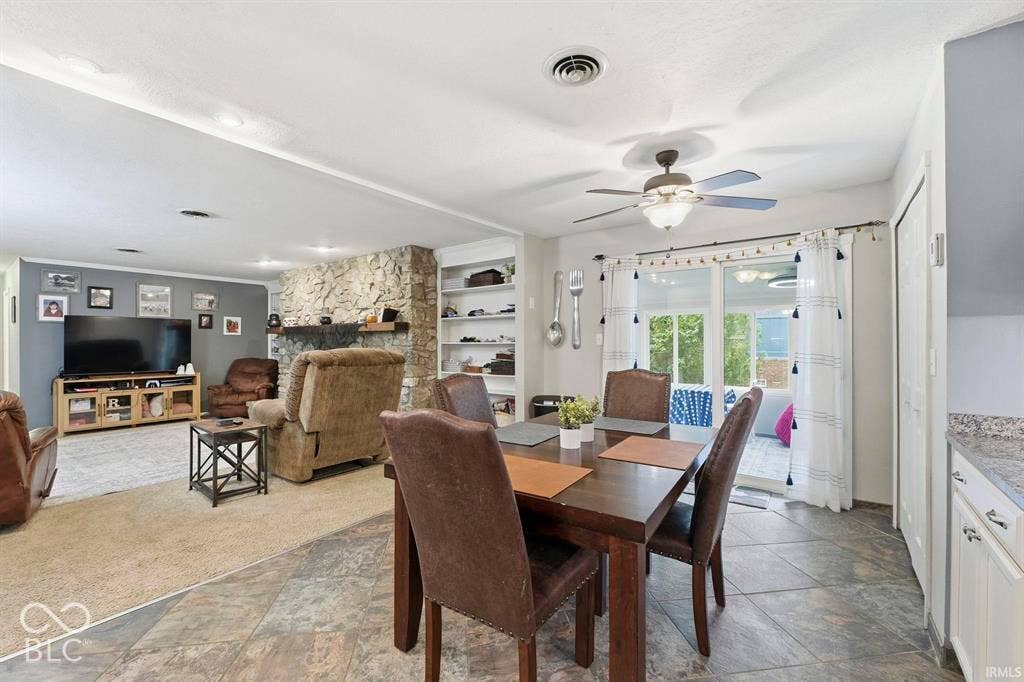 Open-concept living area featuring a wood-finish dining table, tile flooring, recessed and ceiling fan lighting, a gas fireplace with stone surround, built-in shelving, and glass sliding doors leading to outdoor space