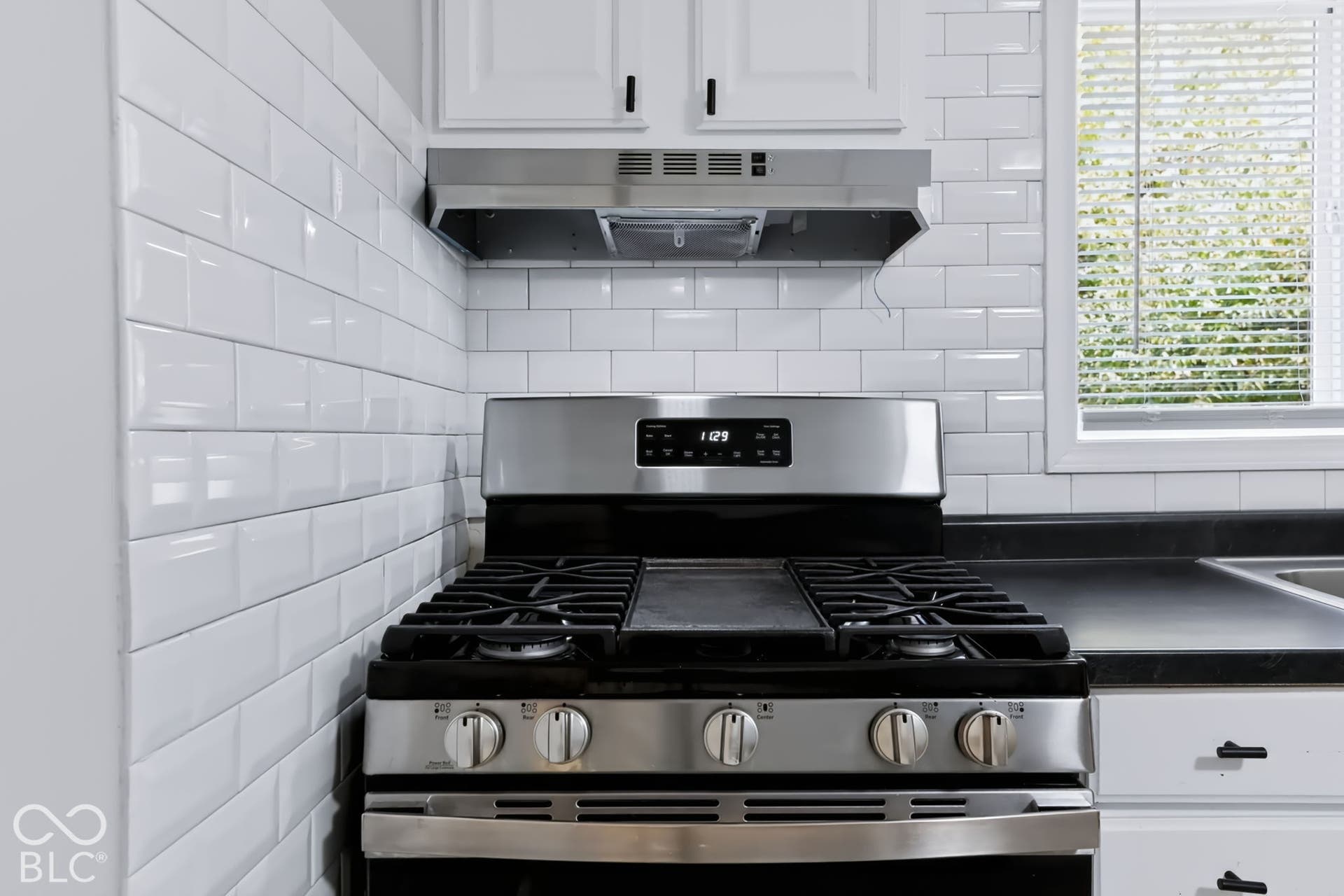 Stainless steel gas range with a coordinating range hood, white subway tile backsplash, white cabinetry with black hardware, and dark countertops
