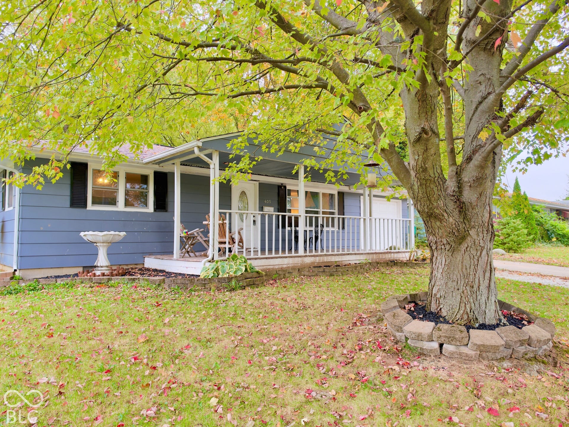 Blue exterior siding featuring a covered front porch with white railing, stone-lined tree bed, and mature tree