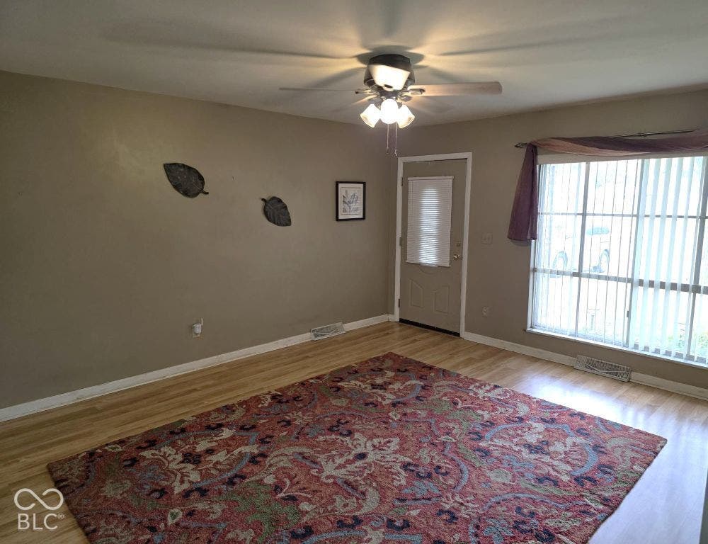 Wood-finish flooring, ceiling fan with integrated lighting, a panel door, and a large window with vertical blinds