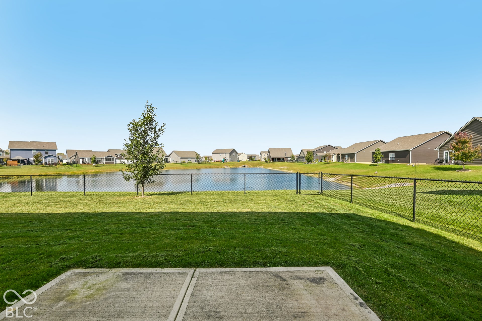 Backyard featuring a concrete patio, grassy lawn, and black chain-link fencing overlooking a pond